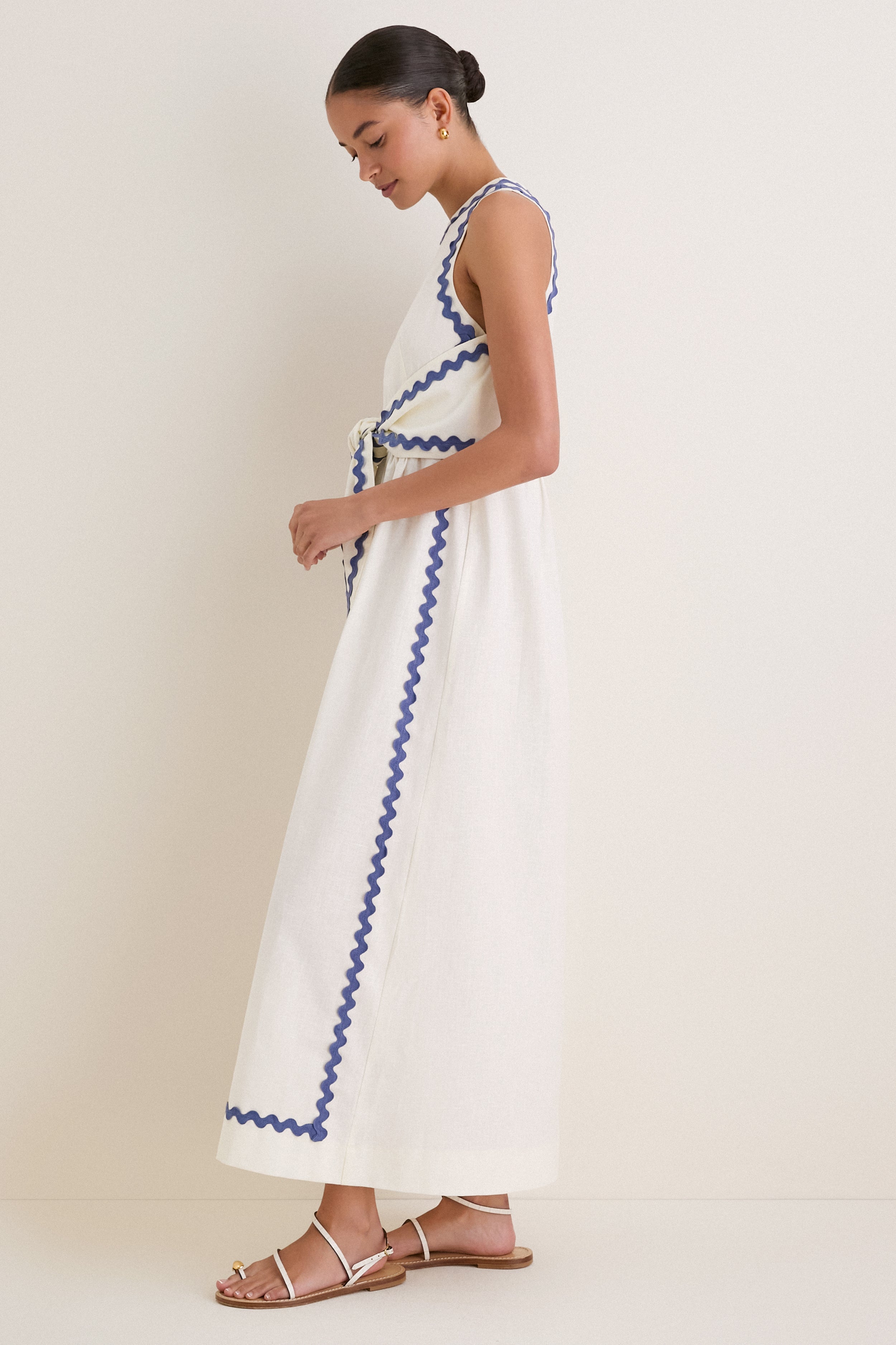 Woman in profile wearing the classic White Ric Rac Tie-Front Brocks Dress, an ankle-length, sleeveless design, exuding effortless elegance with wavy blue ribbon accents.