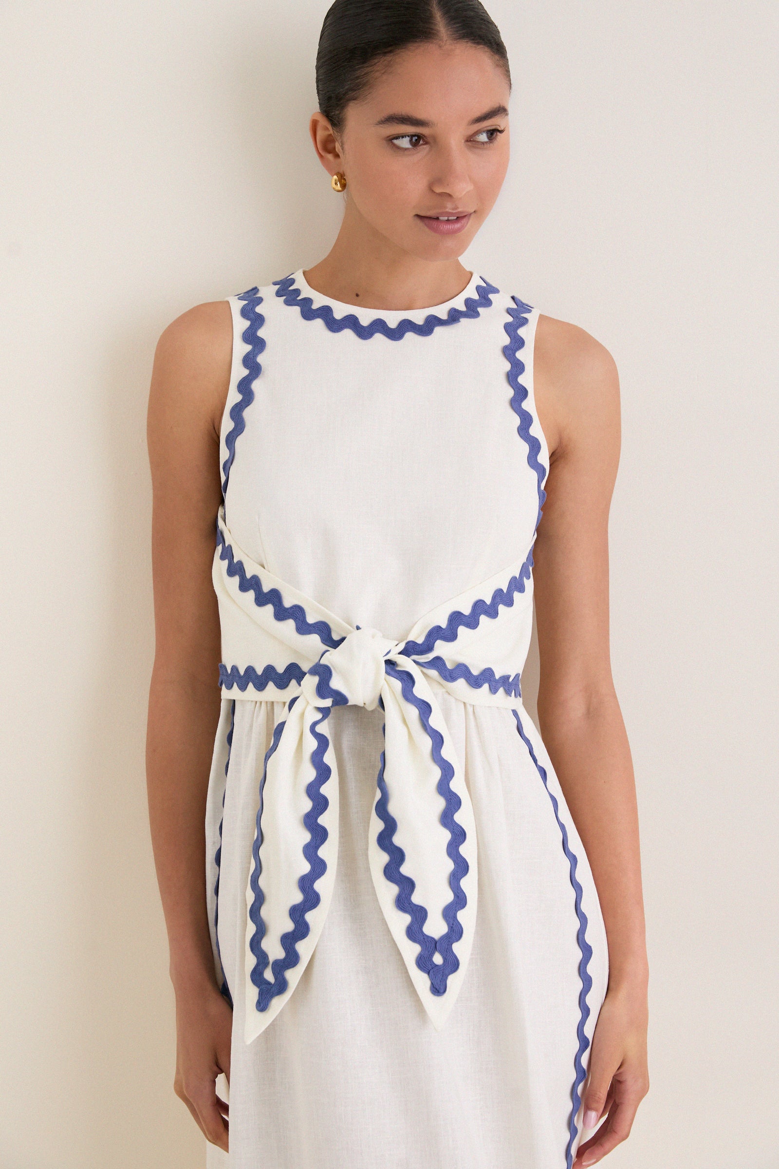 Young woman wearing the classic White Ric Rac Tie-Front Brocks Dress, featuring effortless periwinkle-blue trim and a fun, high-end bow detail at the waist.