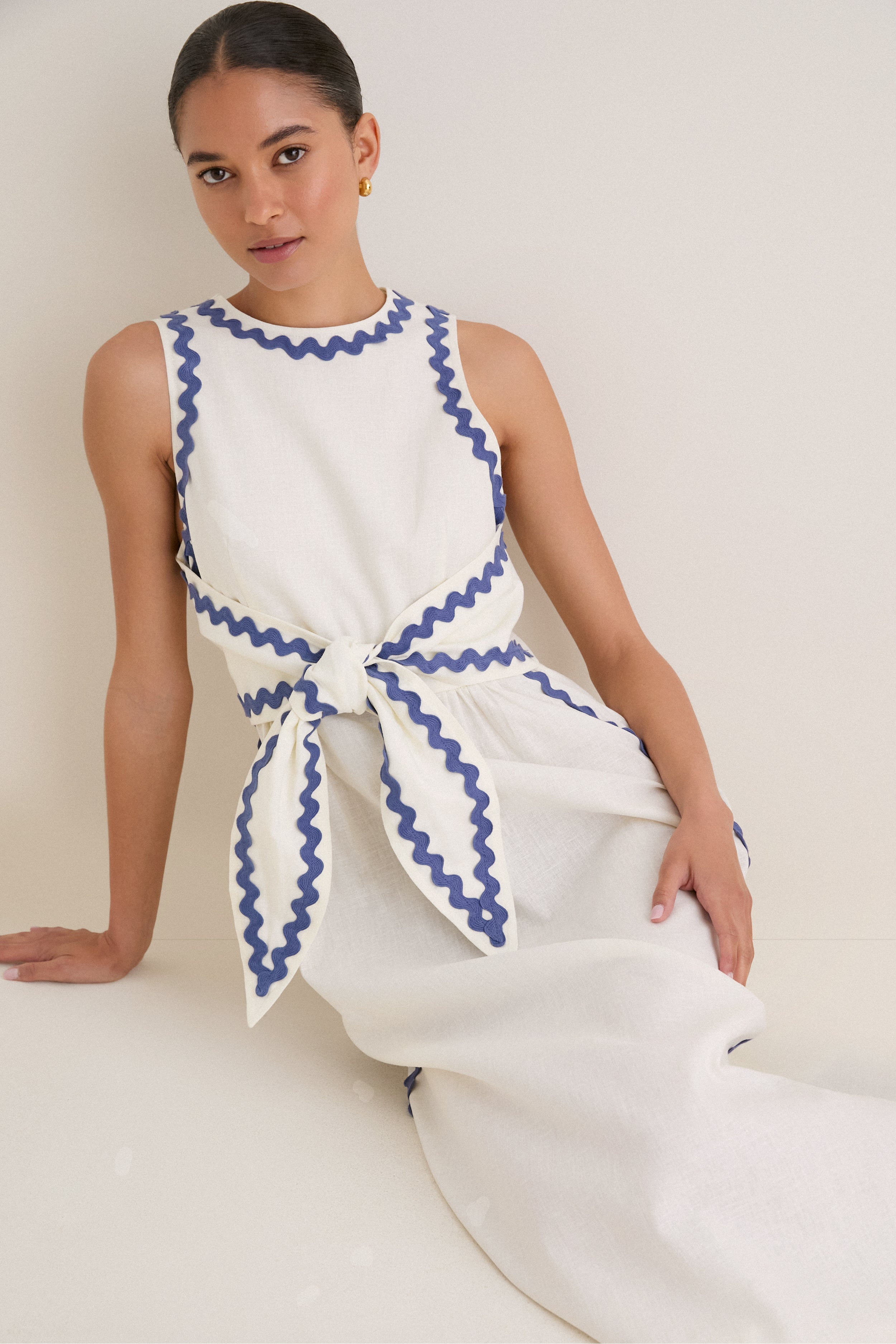 Young woman wearing the classic White Ric Rac Tie-Front Brocks Dress, featuring effortless blue trim details and a fun, high-end sleeveless linen design with a self-tie belt.
