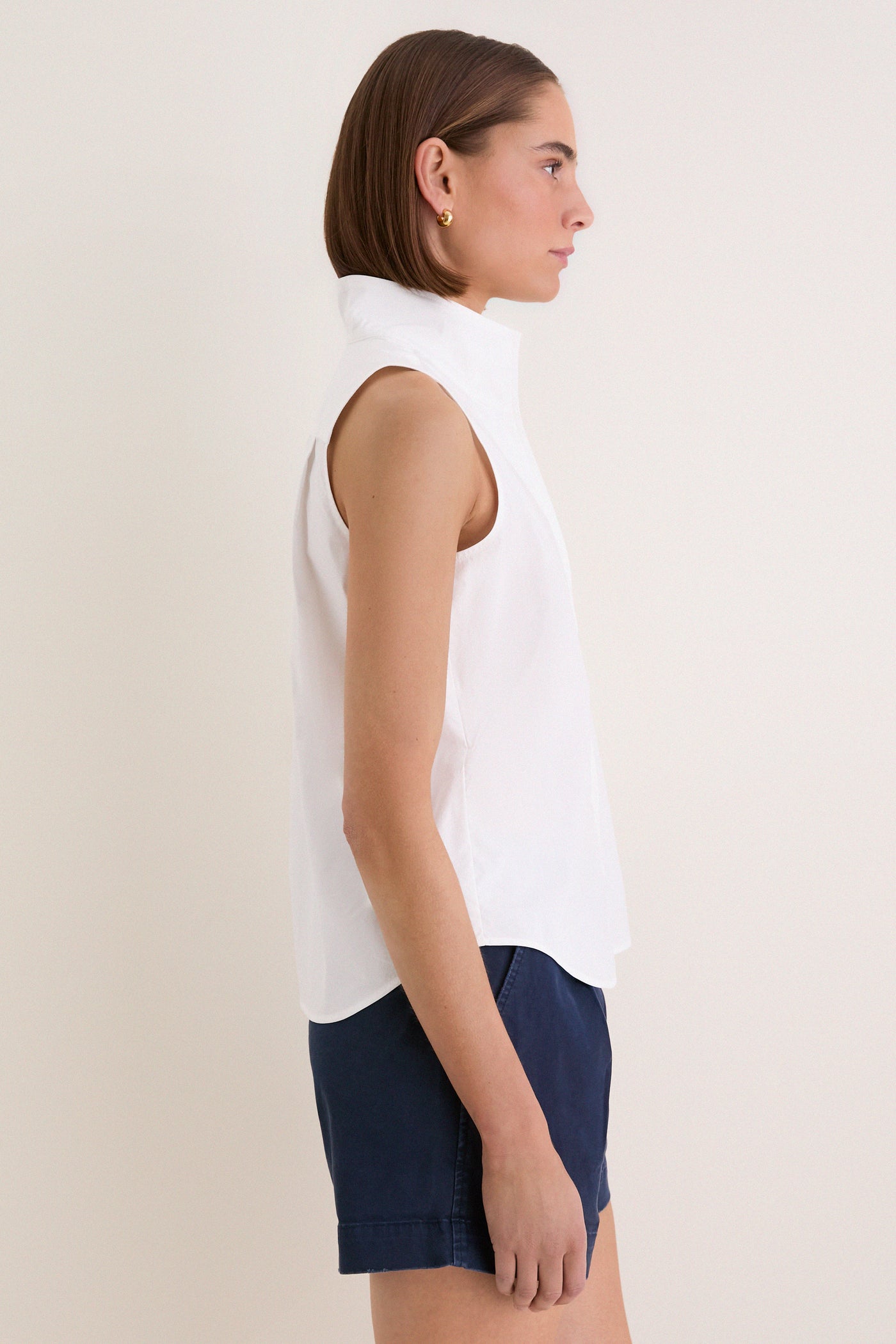 Woman models the classic White Sleeveless Bessette Button Down with stand collar and hidden buttons, paired with navy shorts for an effortless, high-end summer look.