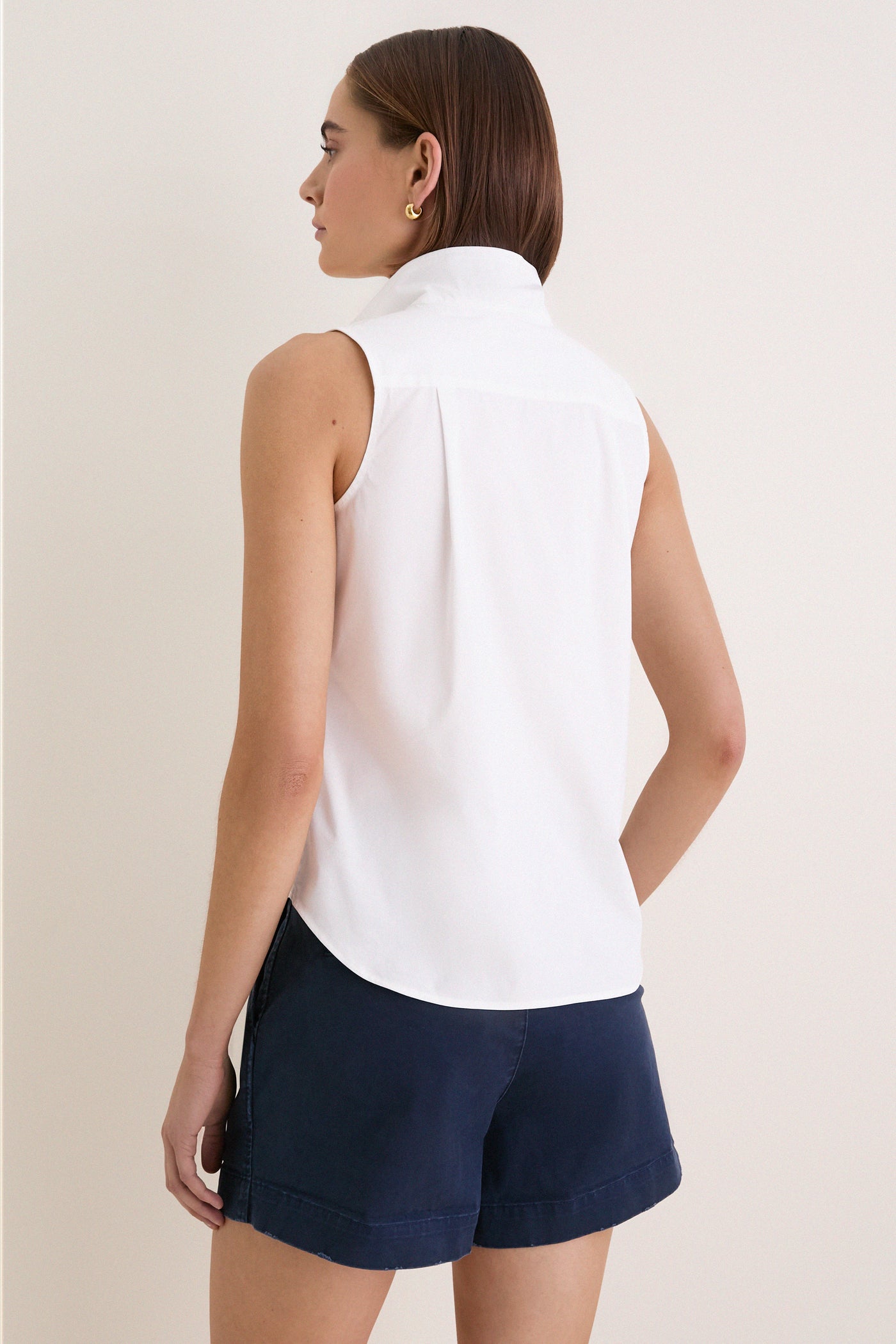 Back view of a woman in a classic, high-end White Sleeveless Bessette Button Down blouse, embodying effortless style with chic details like a stand collar.