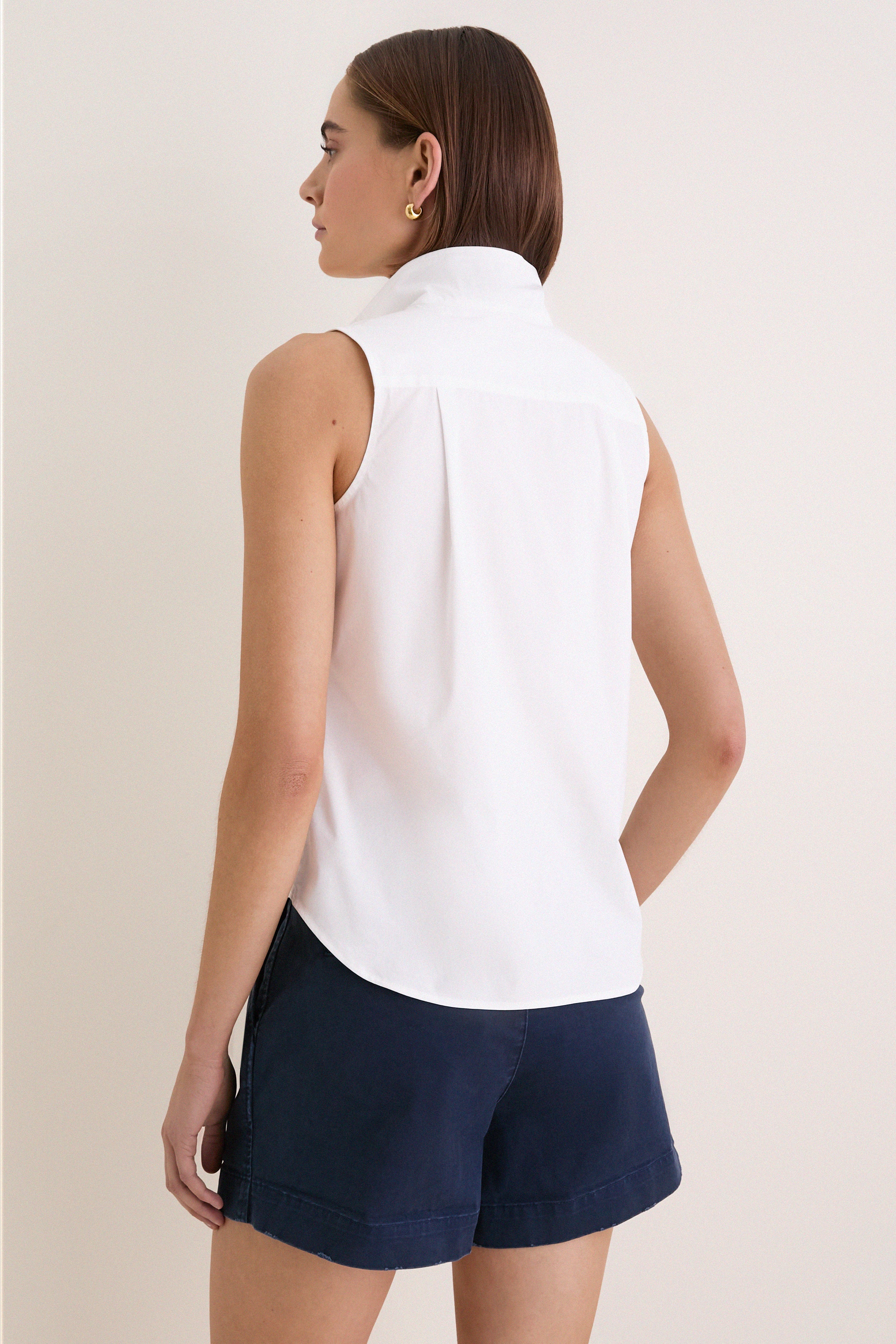 Back view of a woman in a classic, high-end White Sleeveless Bessette Button Down blouse, embodying effortless style with chic details like a stand collar.