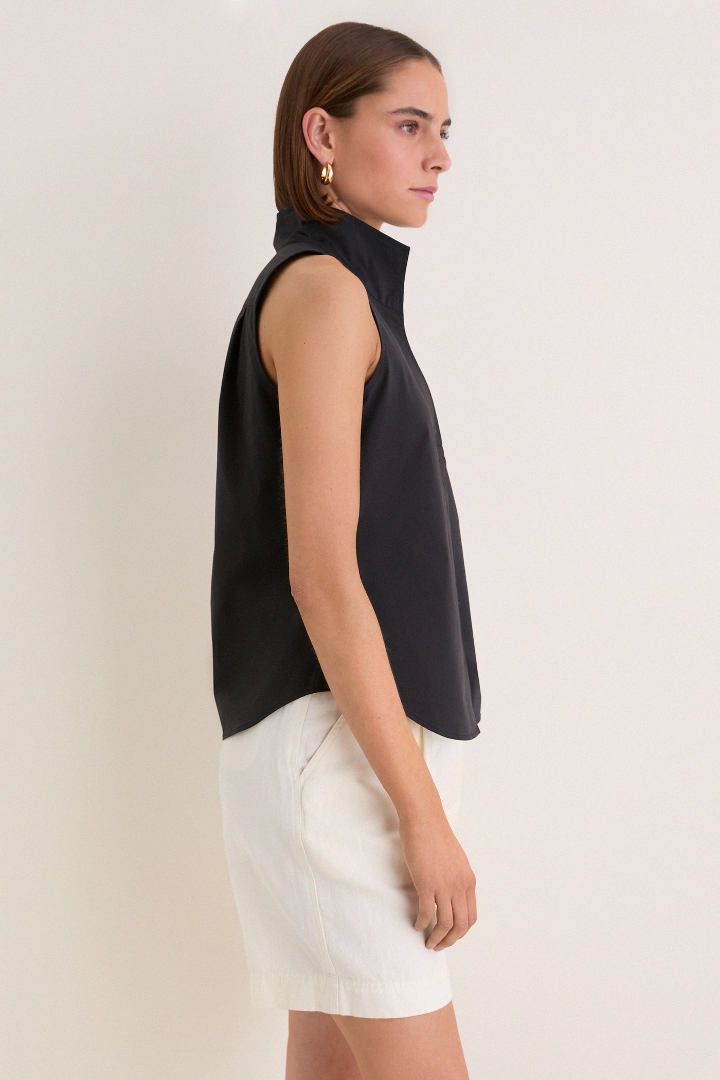 Person models the classic, high-end Black Sleeveless Bessette Button Down with a stand collar, paired with white shorts, showcasing an effortless, fun style.