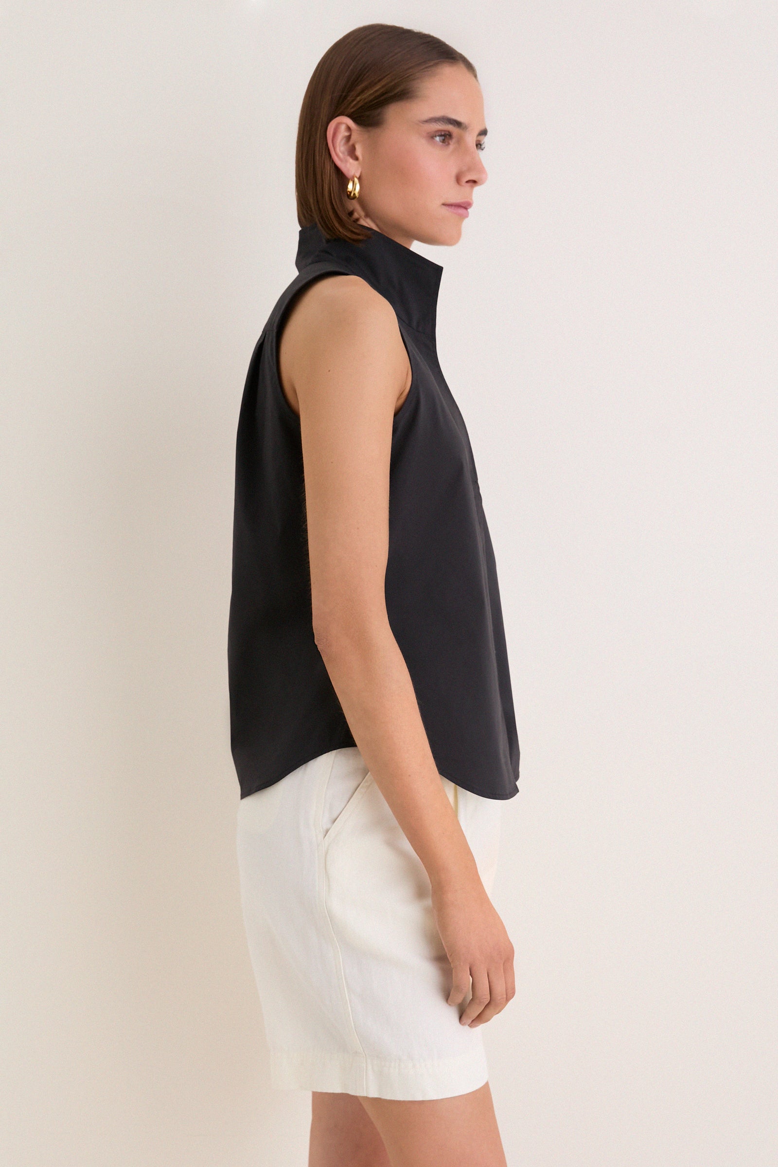 Person models the classic, high-end Black Sleeveless Bessette Button Down with a stand collar, paired with white shorts, showcasing an effortless, fun style.