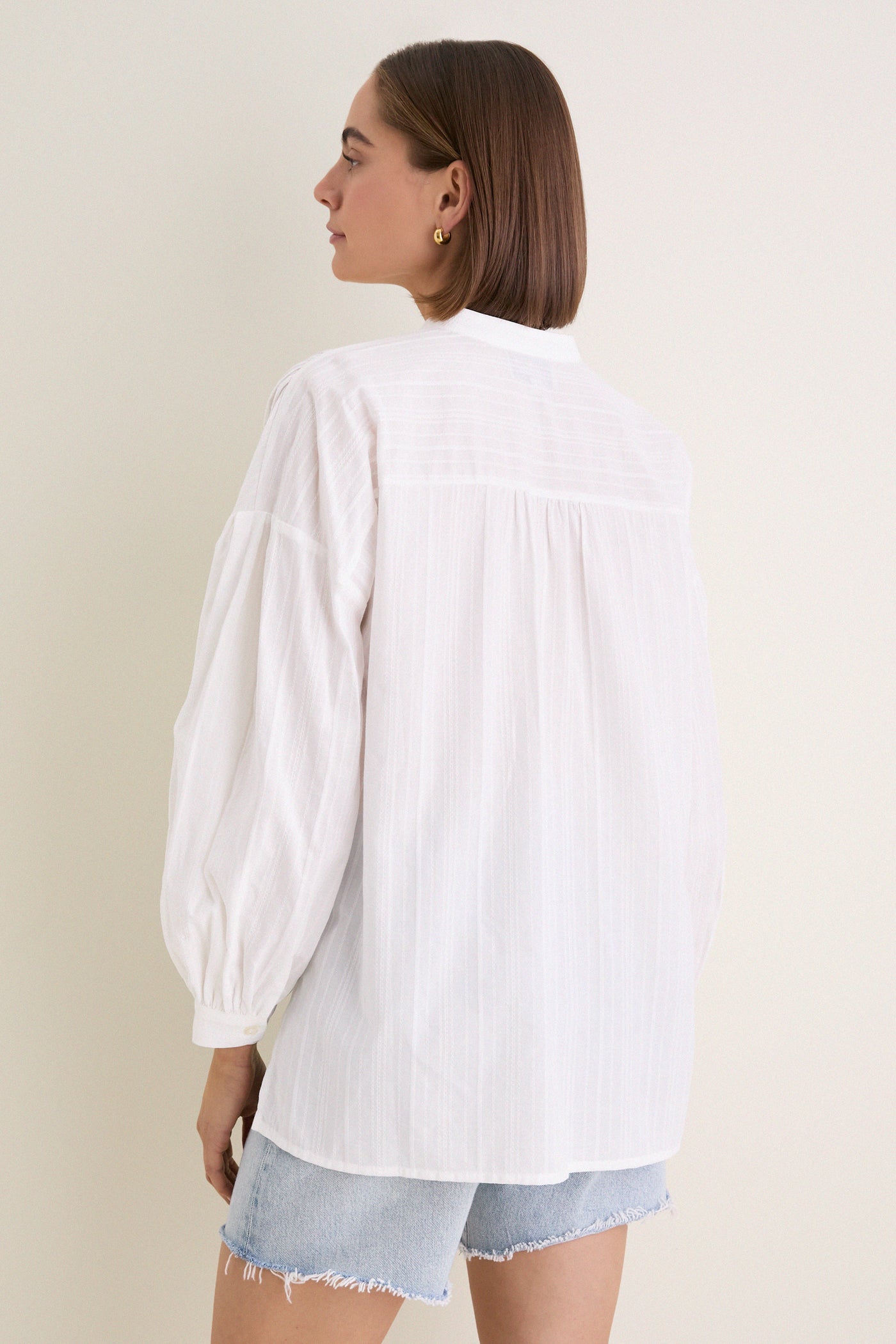 White Shadow Stripe Juliette Button Down with flowy silhouette, gathered sleeves, and asymmetrical hem, worn with denim shorts. Effortless classic style for a fun, high-end look.