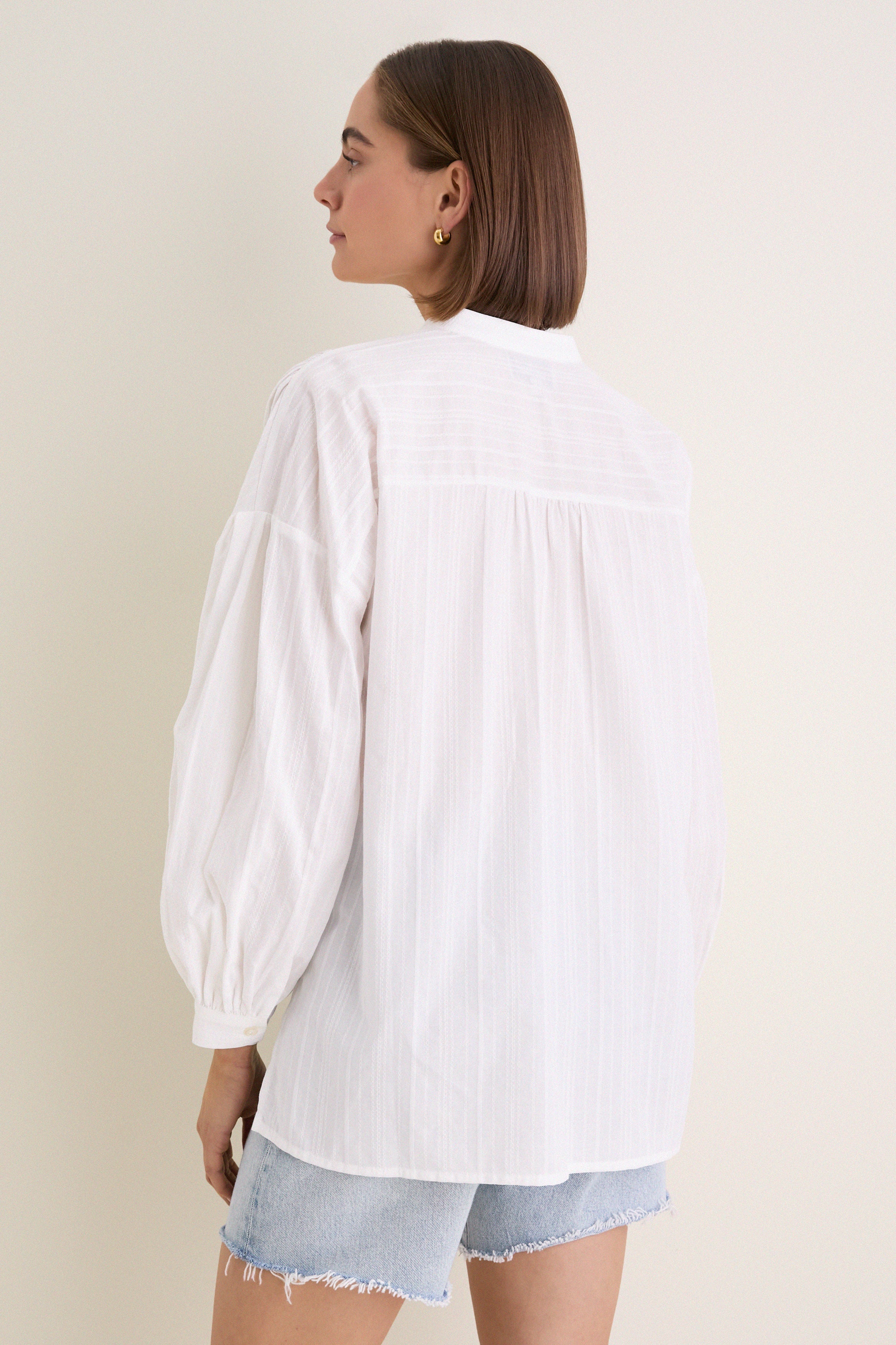White Shadow Stripe Juliette Button Down with flowy silhouette, gathered sleeves, and asymmetrical hem, worn with denim shorts. Effortless classic style for a fun, high-end look.