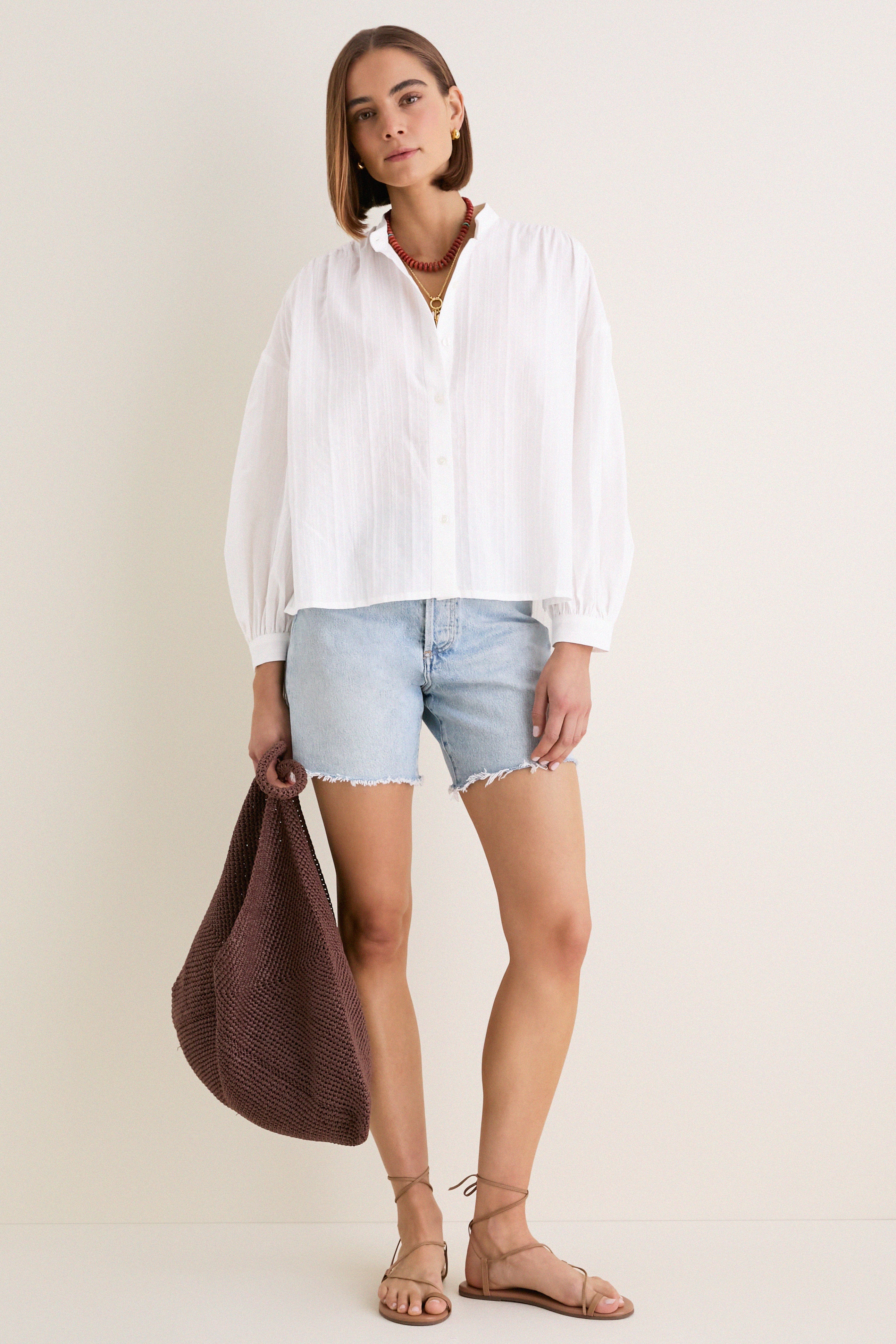 Woman models an effortless, high-end White Shadow Stripe Juliette Button Down with a classic flowy silhouette, paired with frayed denim shorts and strappy sandals.