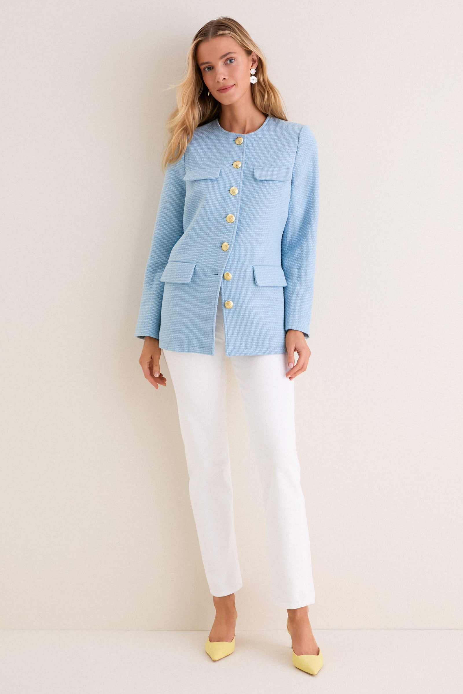 Woman models the Light Blue Tweed Jennings Jacket with gold buttons and flap pockets, styled with white pants and yellow heels for a classic, effortless look.