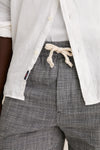 Close-up of India Ink Saturday Beach Short, featuring classic gray-and-white check pattern, elastic waistband with drawstring, over a white tee and open button-down shirt. Effortless and high-end style.