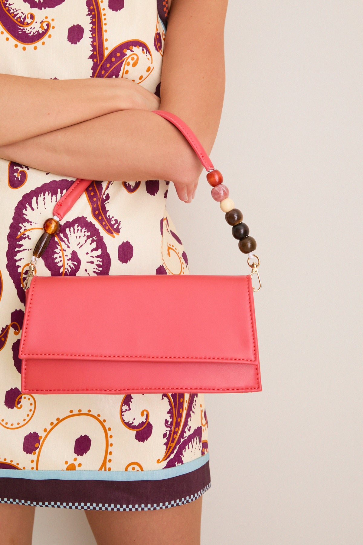 Person holding the Coral Brooks Crossbody, a classic coral-pink envelope handbag with a beaded handle and gold-tone hardware, exuding effortless, high-end style and fun versatility.