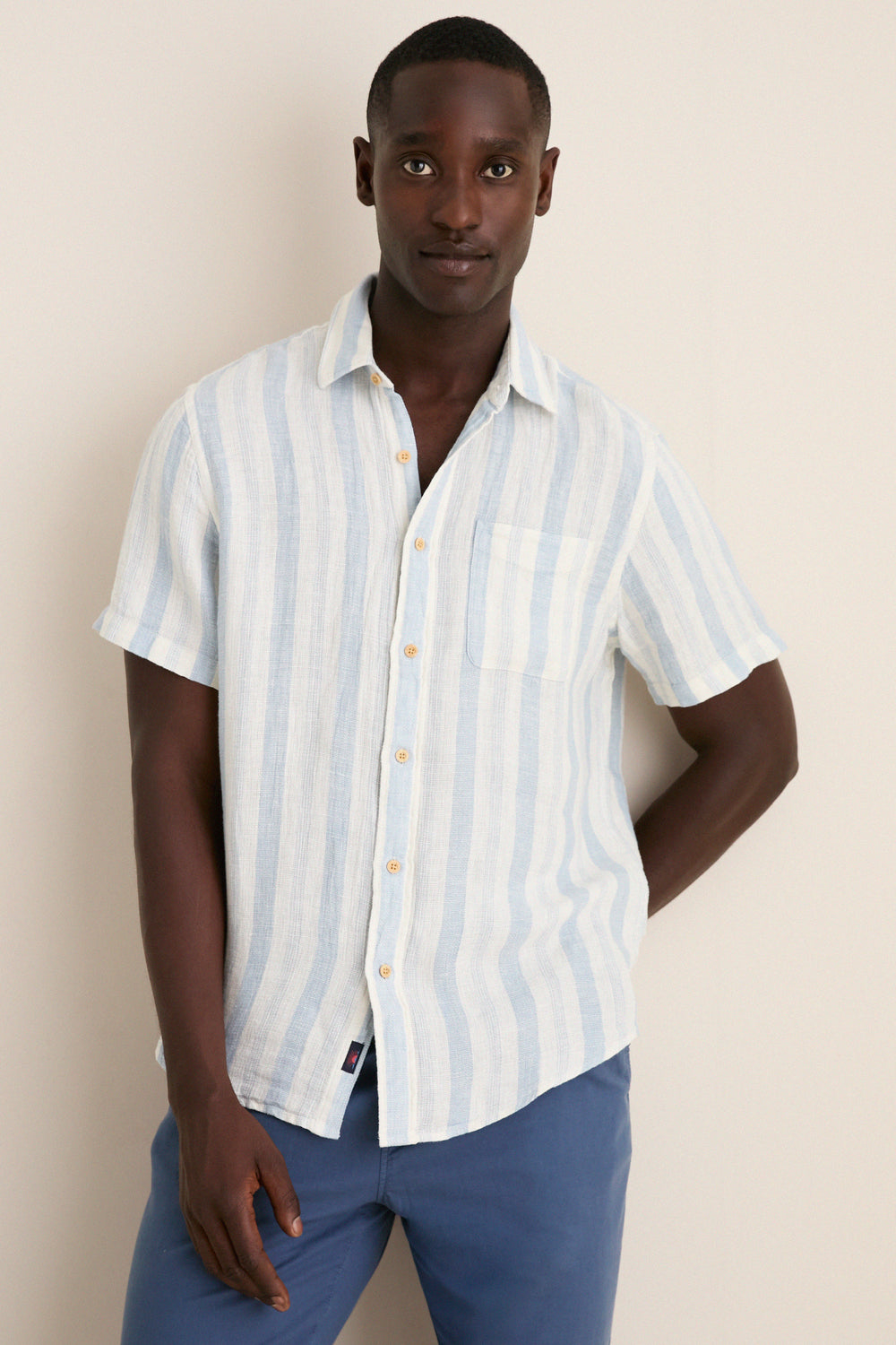 Male model wearing the Island Blue Stripe Palma Linen Shirt, featuring classic pale blue stripes and a chest pocket, paired with medium-blue trousers for an effortless, high-end look.