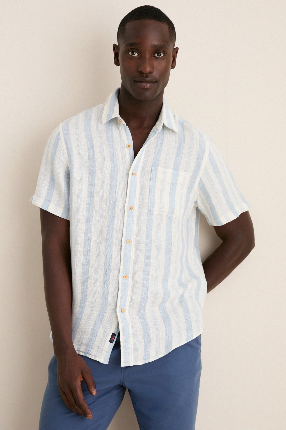 Male model wearing the Island Blue Stripe Palma Linen Shirt, featuring classic pale blue stripes and a chest pocket, paired with medium-blue trousers for an effortless, high-end look.