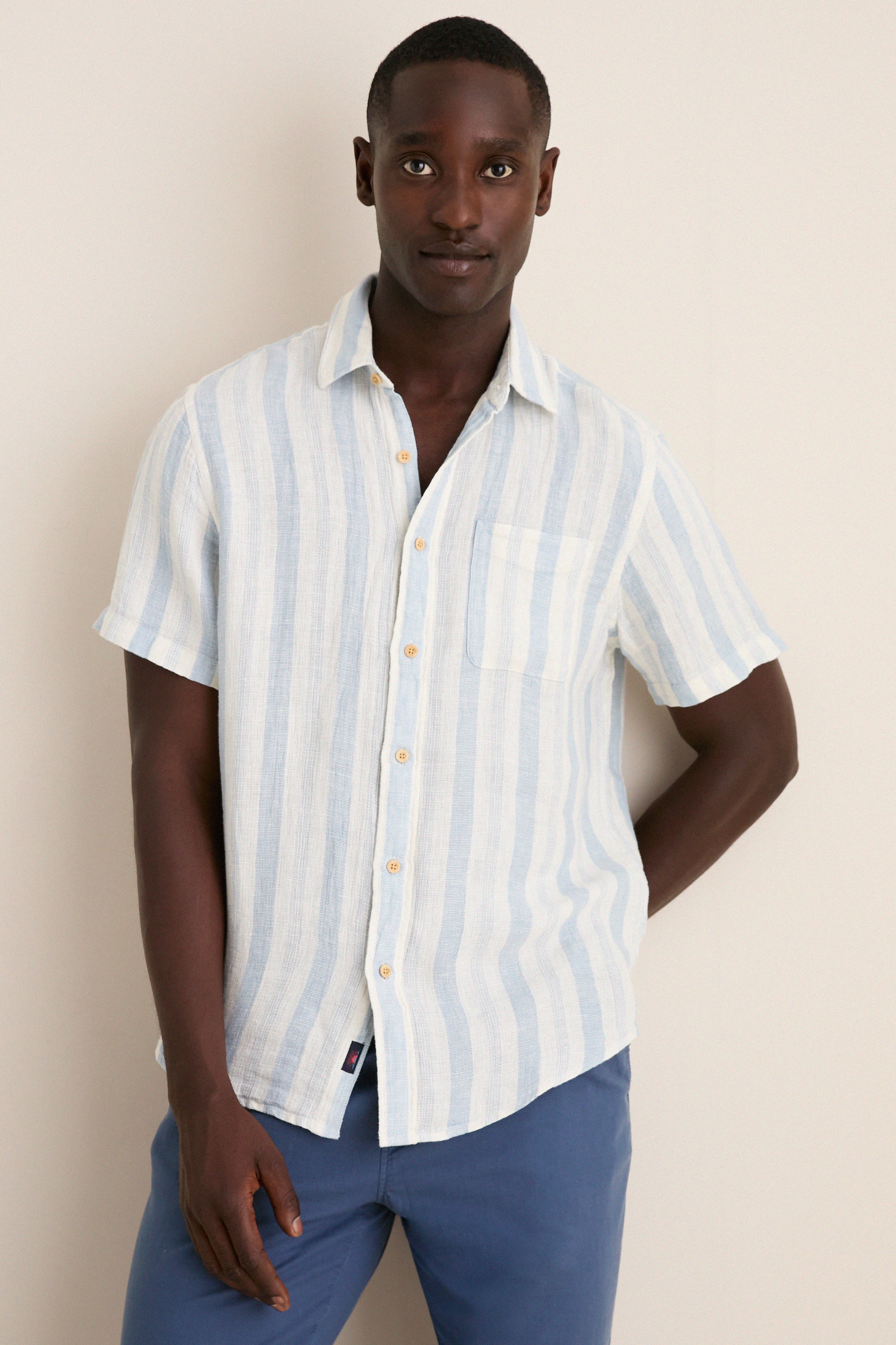 Male model wearing the Island Blue Stripe Palma Linen Shirt, featuring classic pale blue stripes and a chest pocket, paired with medium-blue trousers for an effortless, high-end look.