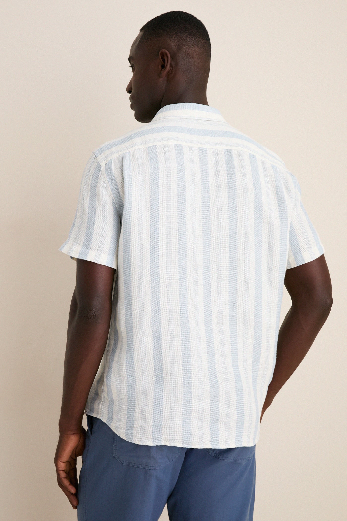 Back view of a man wearing the Island Blue Stripe Palma Linen Shirt, showcasing its classic, effortless style with short sleeves and a relaxed fit, perfect for warm weather.