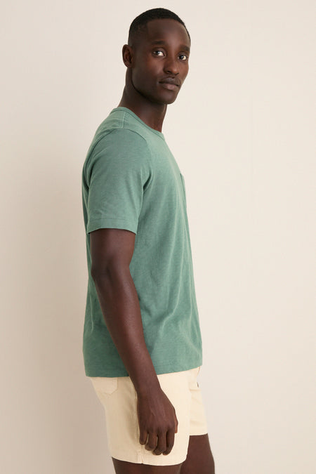 Young man in profile wearing the Island Spruce Sunwashed Pocket Tee, a classic, effortless green T-shirt, styled with light beige shorts for a relaxed, high-end look.