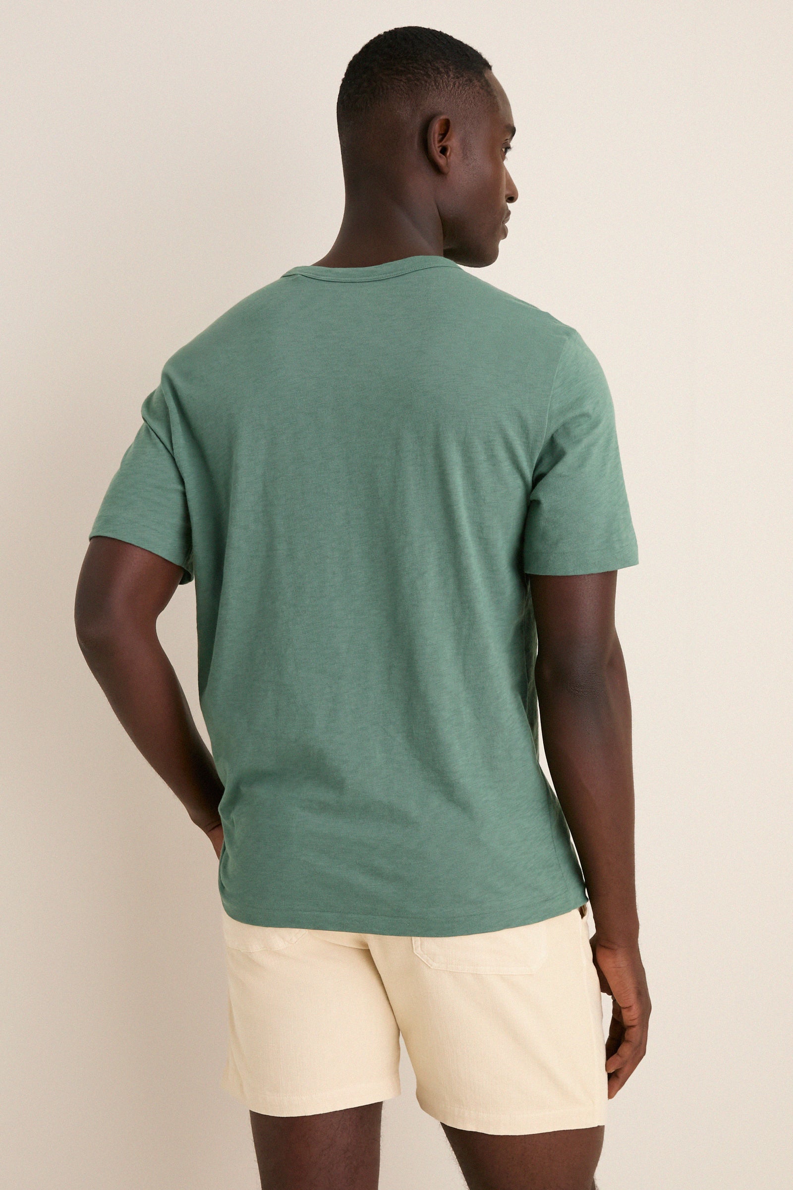 Man wearing the Island Spruce Sunwashed Pocket Tee, showcasing its classic, effortless style with a relaxed fit and pocket detail, paired with off-white shorts.
