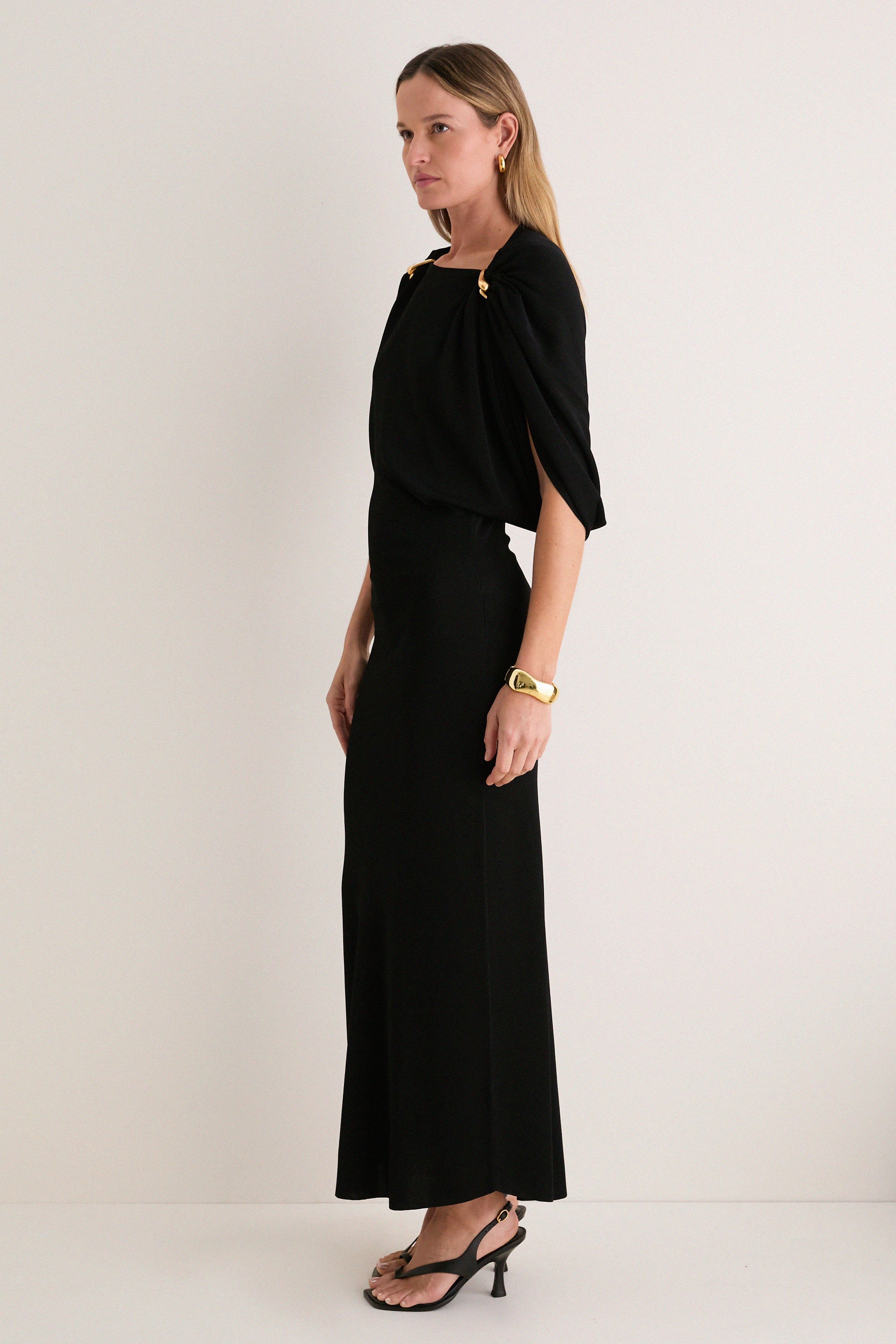 Model in profile wearing the Black Henderson Maxi Dress, featuring classic cape sleeves with gold accents, paired with gold accessories and heels for an effortless, high-end look.