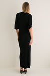 Woman in a Black Henderson Maxi Dress stands effortlessly against an off-white wall, showcasing classic elbow-length sleeves and high-end style with fun, strappy sandals.