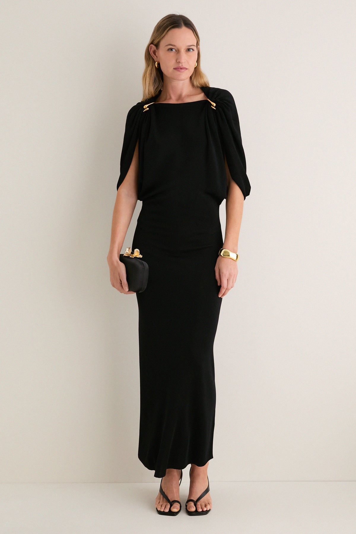 Woman in a classic Black Henderson Maxi Dress with cape sleeves, holding a clutch, accessorized with gold jewelry; an effortless, high-end look for any occasion.