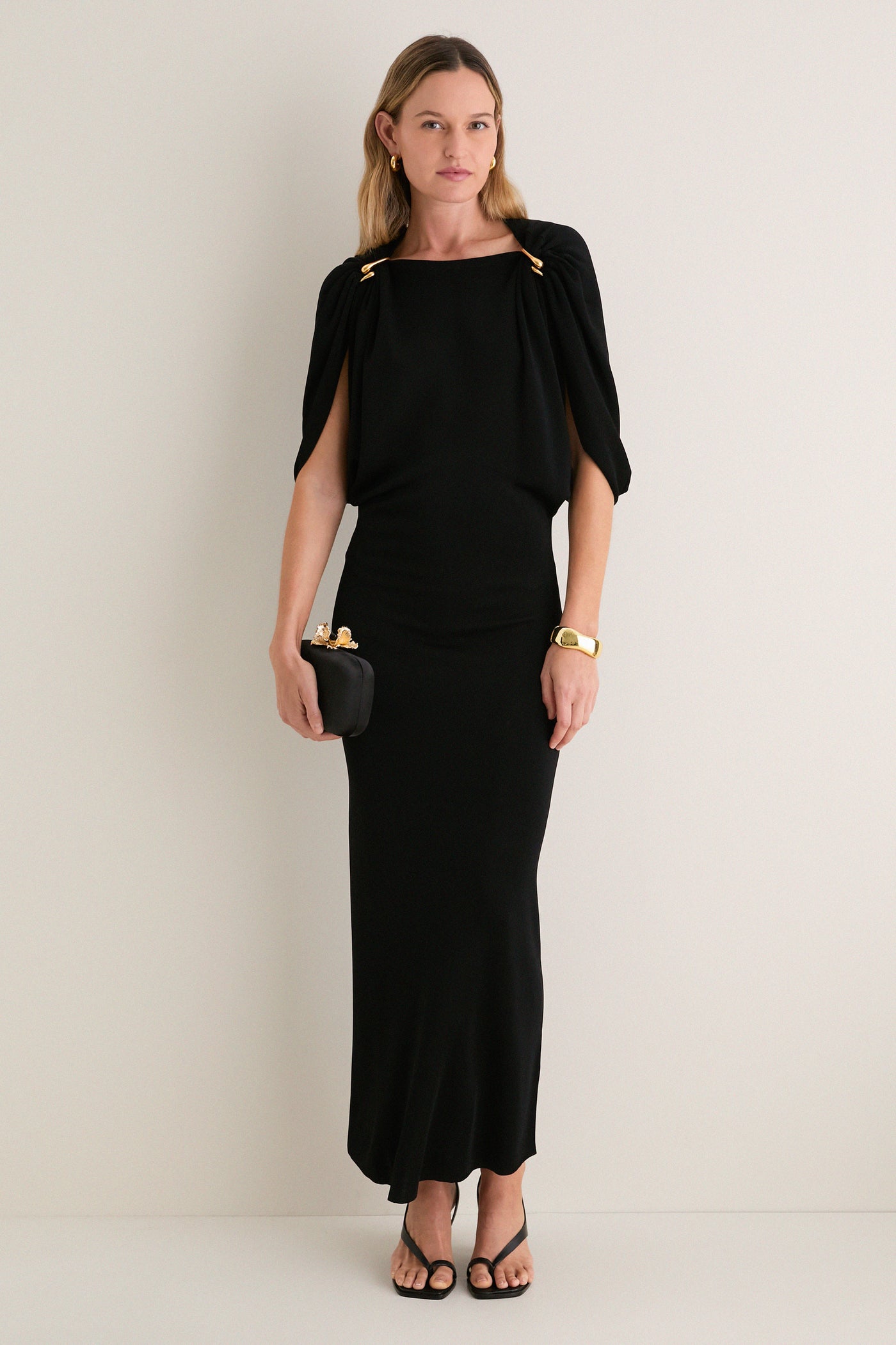 Woman in a classic Black Henderson Maxi Dress with cape sleeves, holding a clutch, accessorized with gold jewelry; an effortless, high-end look for any occasion.
