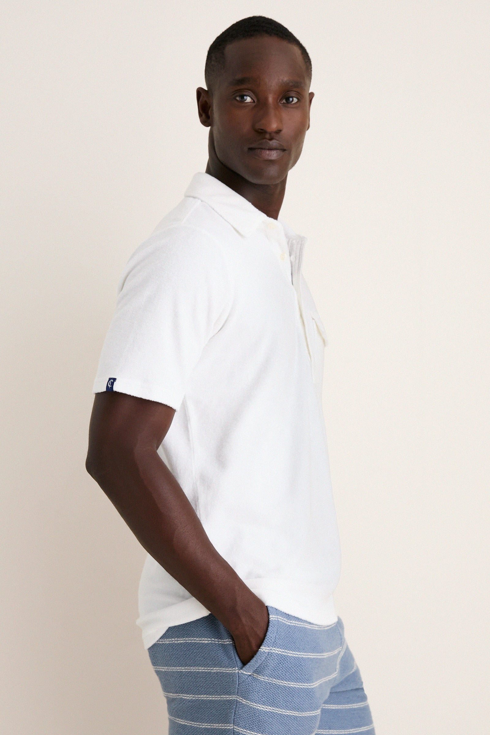 Young man wearing the classic White Lotus Terrycloth Players Shirt, effortlessly styled with light blue striped shorts, embodying a fun, high-end summery look.