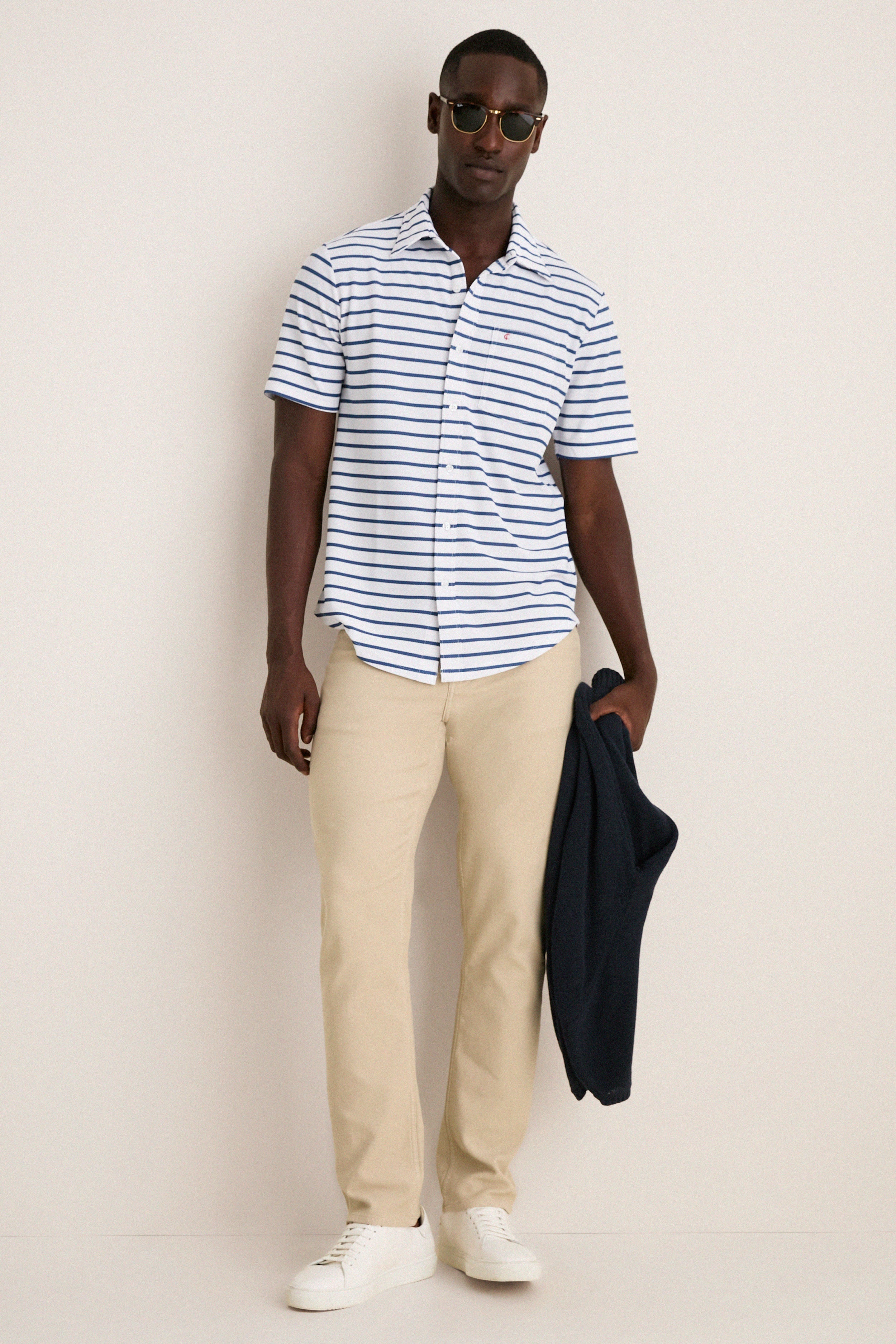 Man wearing the classic White Performance Pique Knit Button Down with navy stripes, khakis, and sneakers, holding a navy jacket. Effortlessly high-end and perfect for fun, warm days.