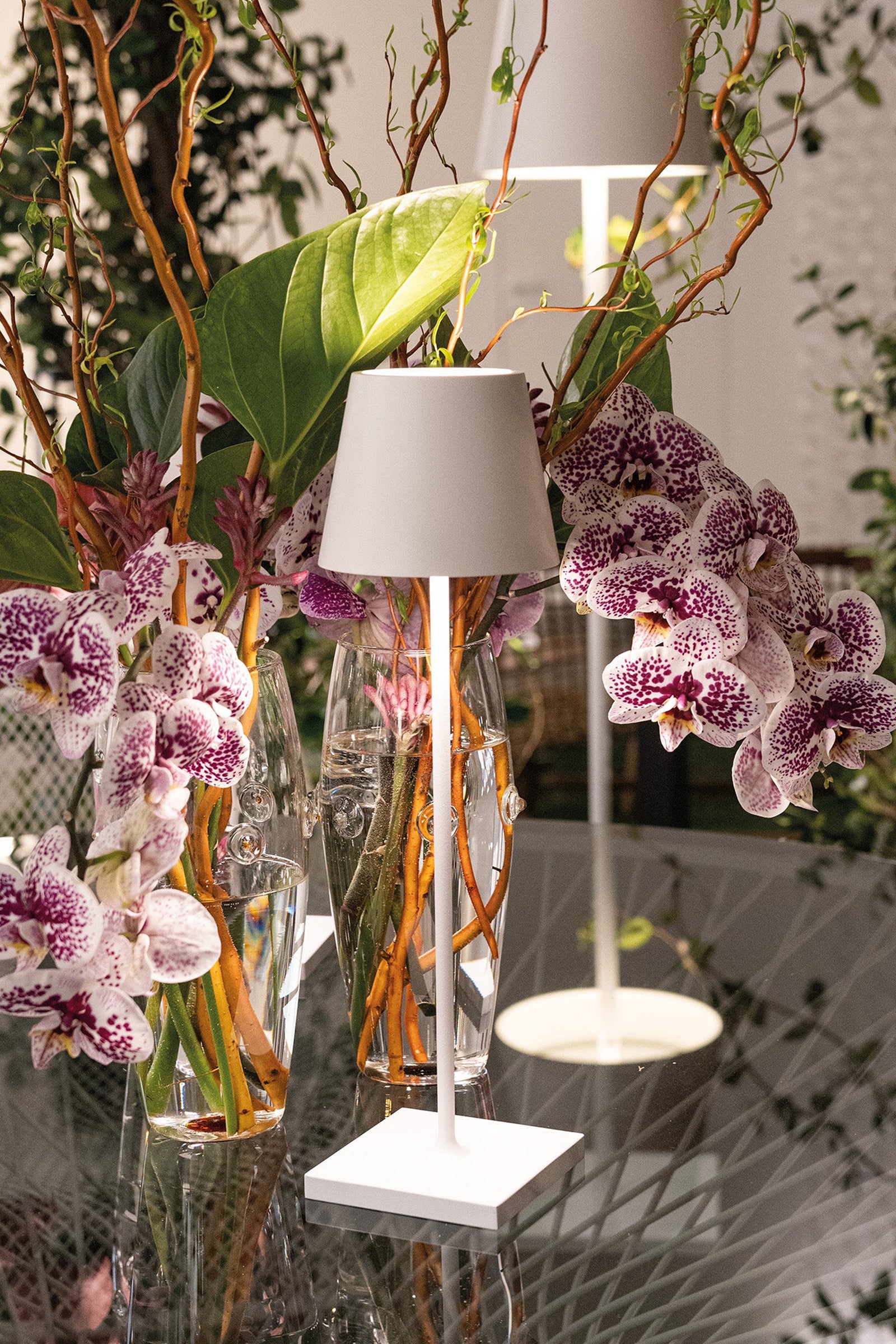 White Poldina Pro Table Micro Lamp illuminates elegantly with a cordless, dimmable design, paired with orchids and foliage in glass vases on a reflective surface.