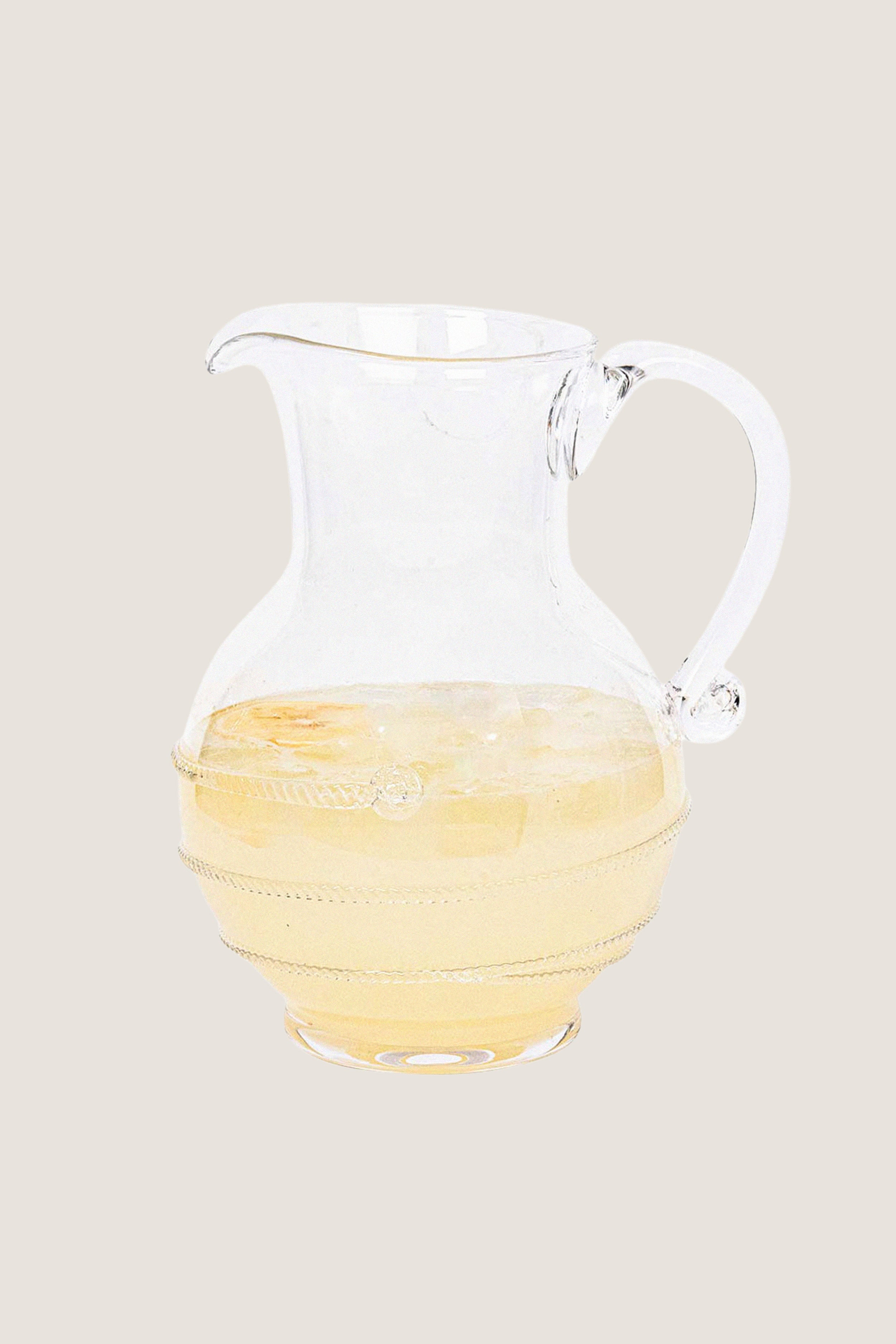 Amalia Glass Round Pitcher, a classic, high-end, mouth-blown glass jug with a swirling thread and berry design, half-filled with a light-colored drink over ice, embodying effortless elegance.