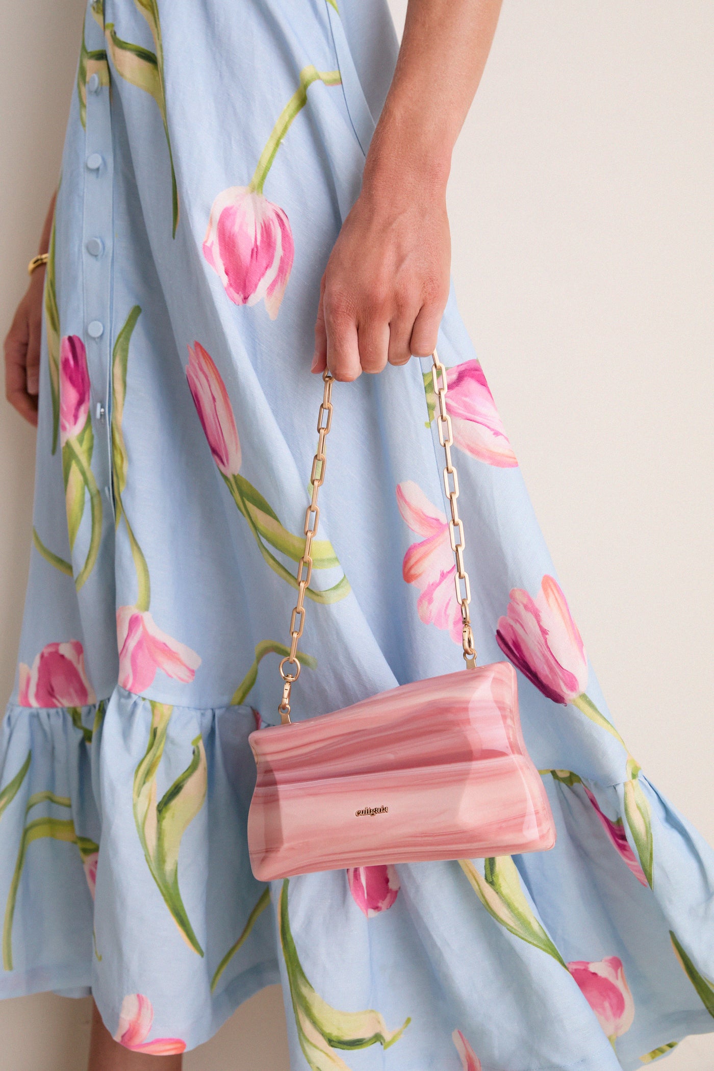 Person holding Rose Water Malaya Clutch, an asymmetrical, marbled acrylic bag with gold-link chain, paired with a classic floral dress for an effortless, high-end look.