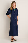 Young woman in a classic, navy Vincent Dress with button-front and puffed sleeves, exuding effortless style, complemented by a bold green pendant and dark brown sandals.