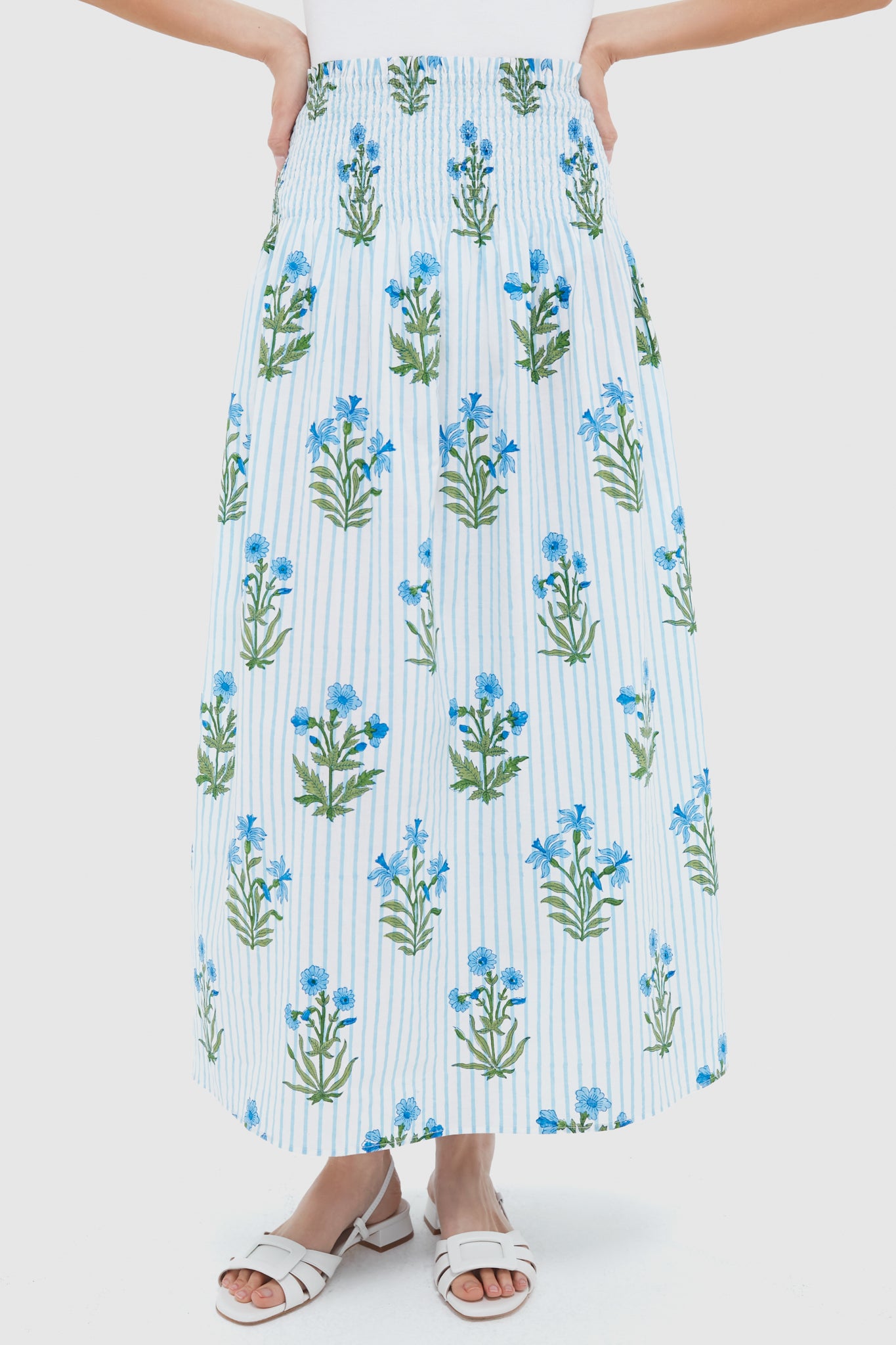 A person wears a Pink City Prints long, white block-print skirt with blue floral and green leaf designs, vertical light blue stripes, and white sandals. Handmade from organic cotton in India for unique style.