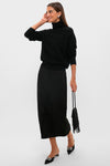 A woman in a Tuckernuck black turtleneck sweater, black suede midi skirt, black pointed-toe shoes with white details, and holding a black fringe handbag.