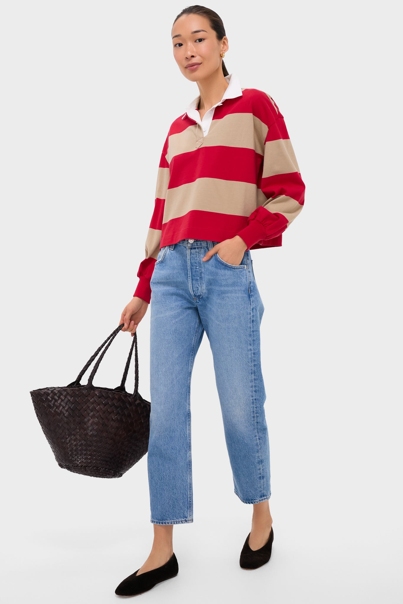 A woman holds a black woven tote bag from Tuckernuck, wearing a ruby striped polo, light blue jeans, and black flats.
