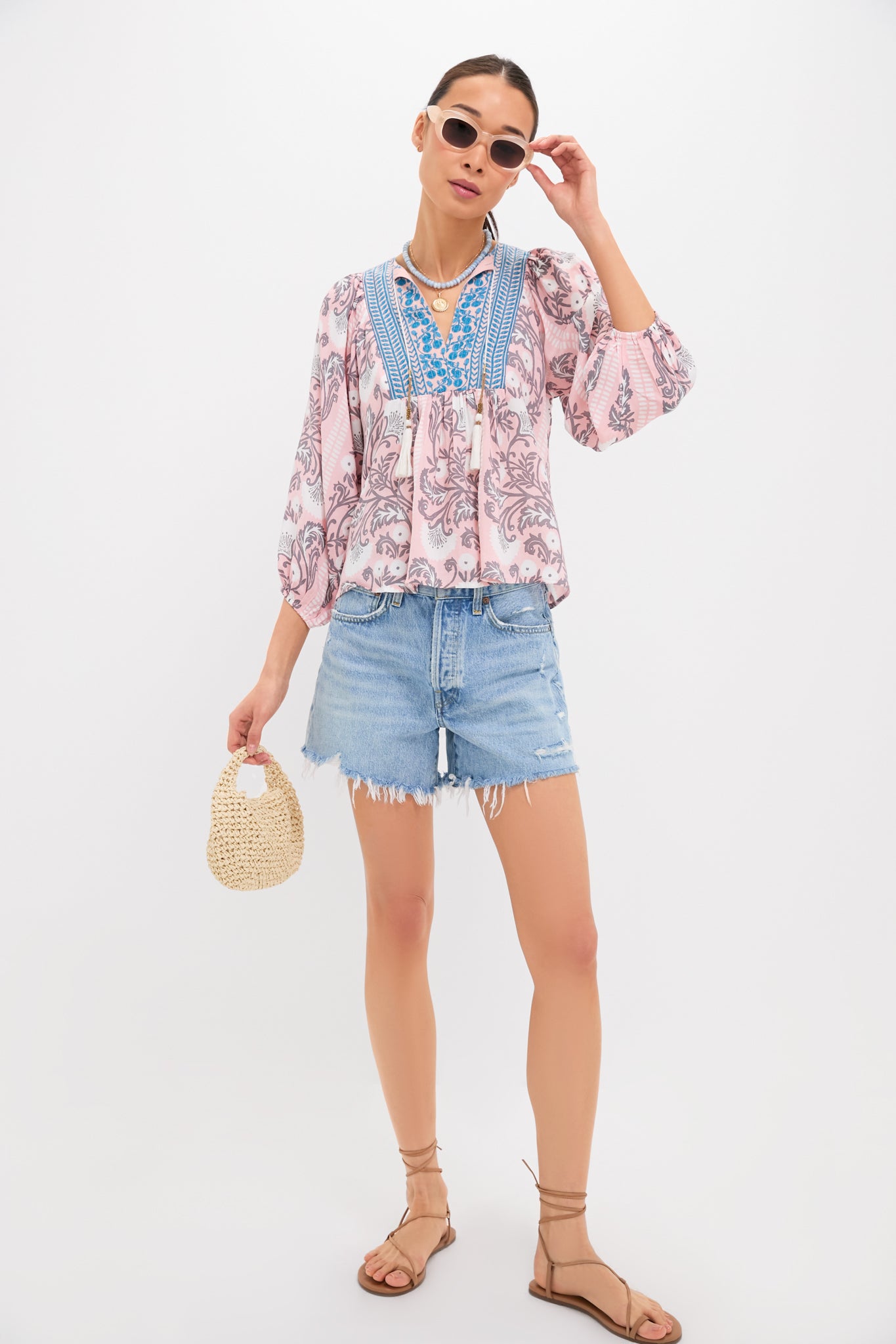 A woman models Bell by Alicia Bell: sunglasses, a boho blouse with printed yoke and blue embroidery, distressed denim shorts, strappy sandals, and a small woven handbag.