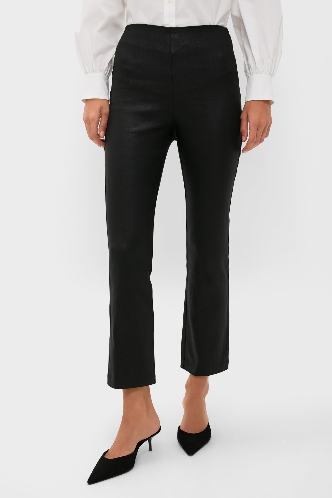 A person wears Tuckernuck Black Wax Ashford Pants, a white long-sleeve blouse, and black pointed-toe heeled mules.