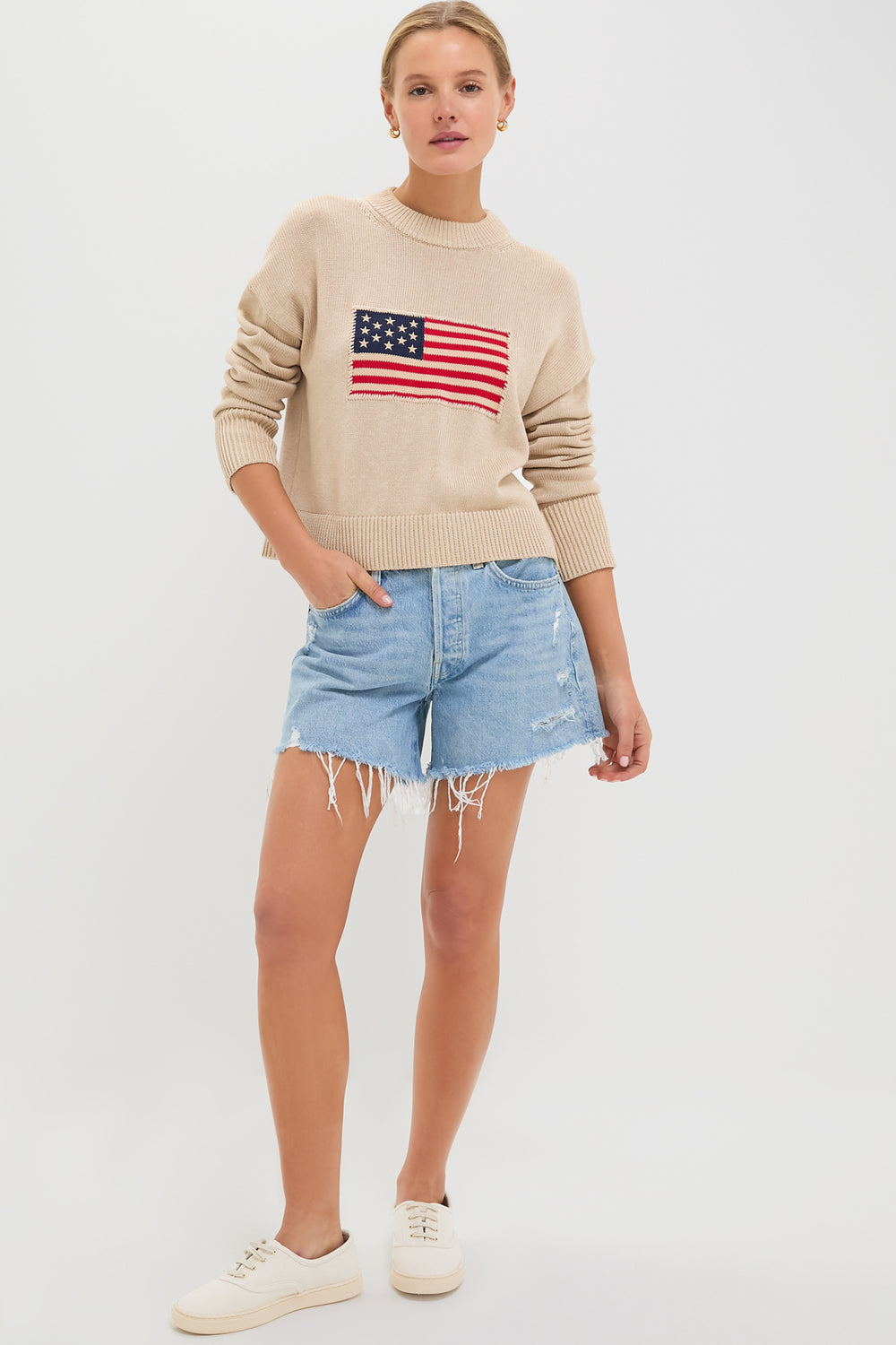 A woman wears a Tuckernuck Cropped Americana Sweater with an intarsia flag, distressed denim shorts, and white sneakers. The sweater’s boxy fit gives her casual look a modern edge.