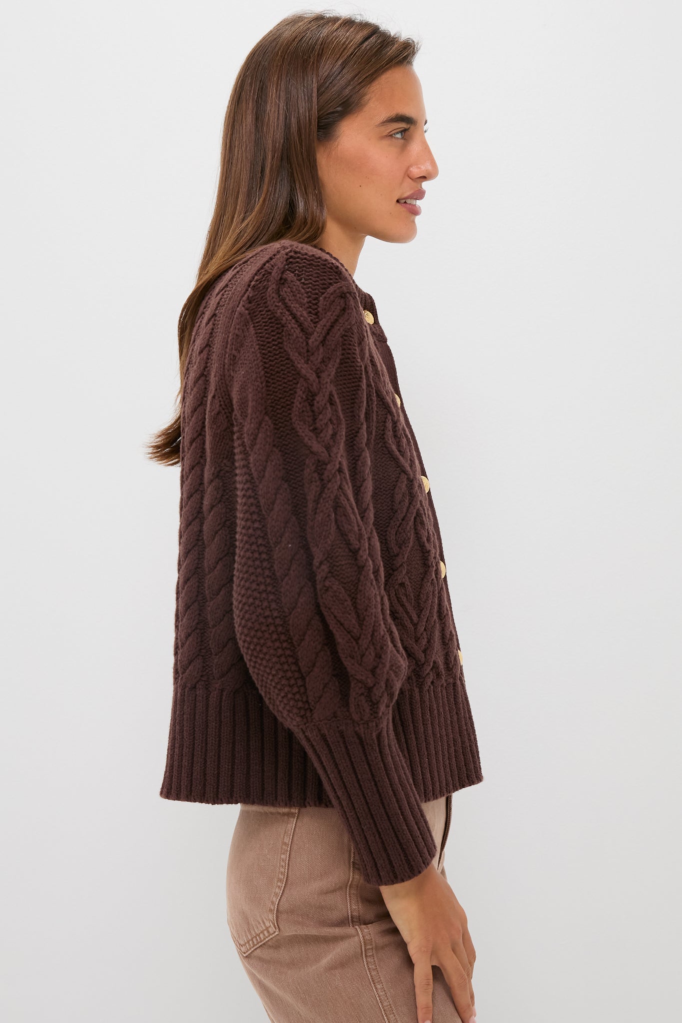 A woman stands in profile against a plain white background, wearing a cropped cable-knit Tuckernuck Colette Cardigan with large buttons and light brown pants.