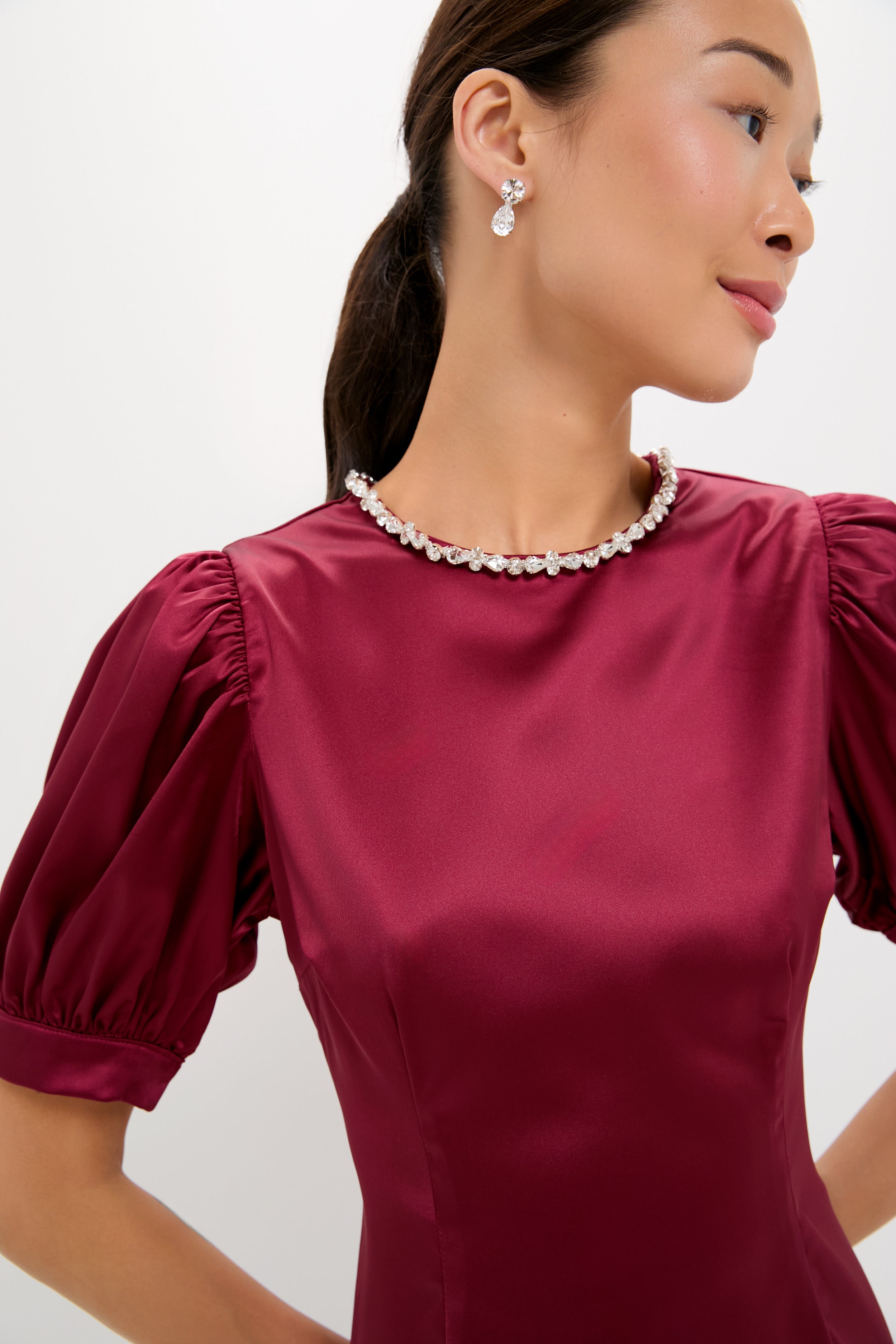 A woman in a shiny burgundy Hyacinth House fit-and-flare dress with puffed sleeves, pearl necklace, and drop earrings stands slightly turned against a plain light background.