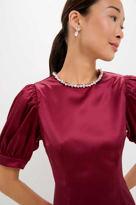 A woman in a shiny burgundy Hyacinth House fit-and-flare dress with puffed sleeves, pearl necklace, and drop earrings stands slightly turned against a plain light background.