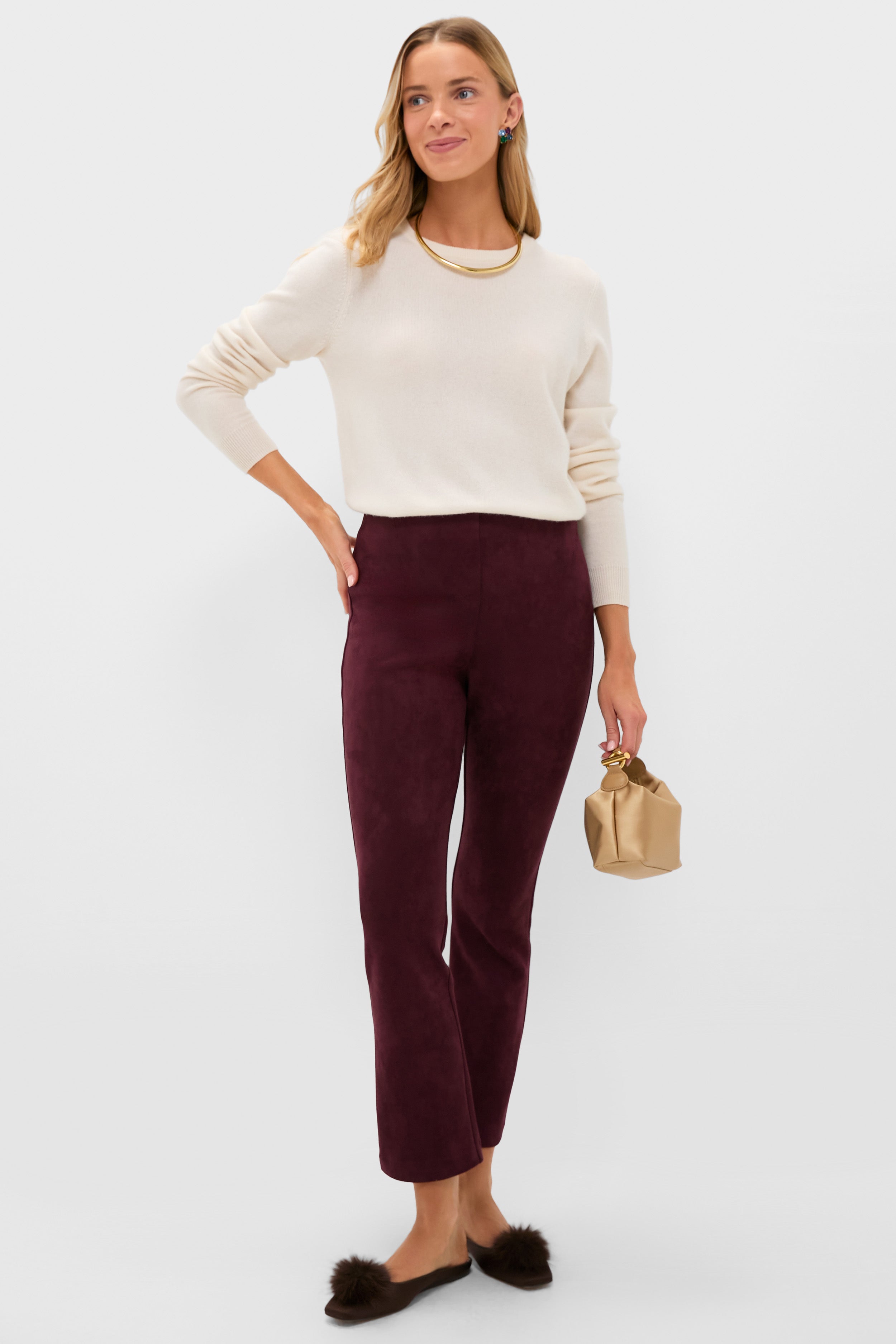 A woman stands against a plain background wearing a Tuckernuck ivory cashmere crewneck sweater, burgundy pants, black slip-on shoes, and holding a small tan pouch.