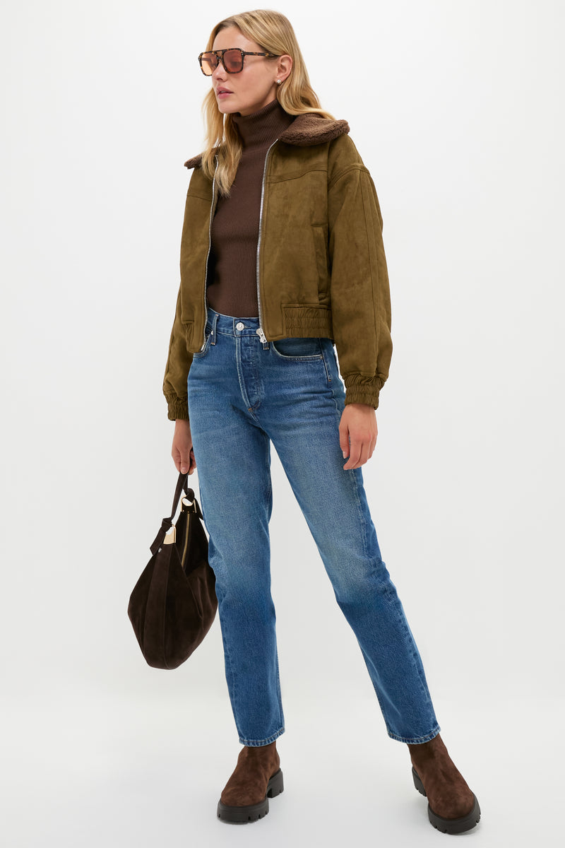 A woman wearing sunglasses, a brown jacket over a brown turtleneck, blue jeans, and brown boots holds a dark brown Stuart Weitzman handbag while standing against a plain background.