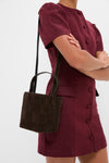 Person wearing a classic burgundy dress holds the Woven Tmoro Suede Laith Top Handle Bag, showcasing its effortless, high-end patchwork design and fun, versatile drawstring closure.
