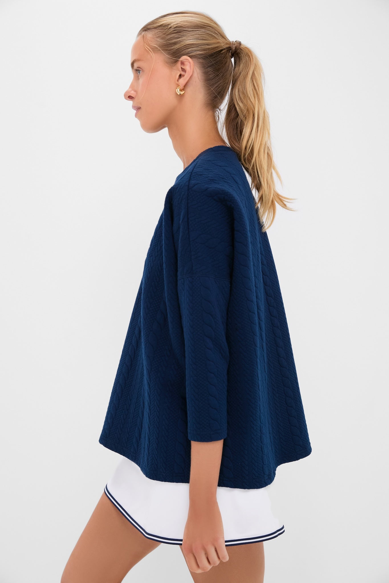 Young woman models the Navy Cable Ally Swing Sweatshirt, showcasing its classic, relaxed fit and quilted texture, paired effortlessly with a fun, high-end tennis skirt.