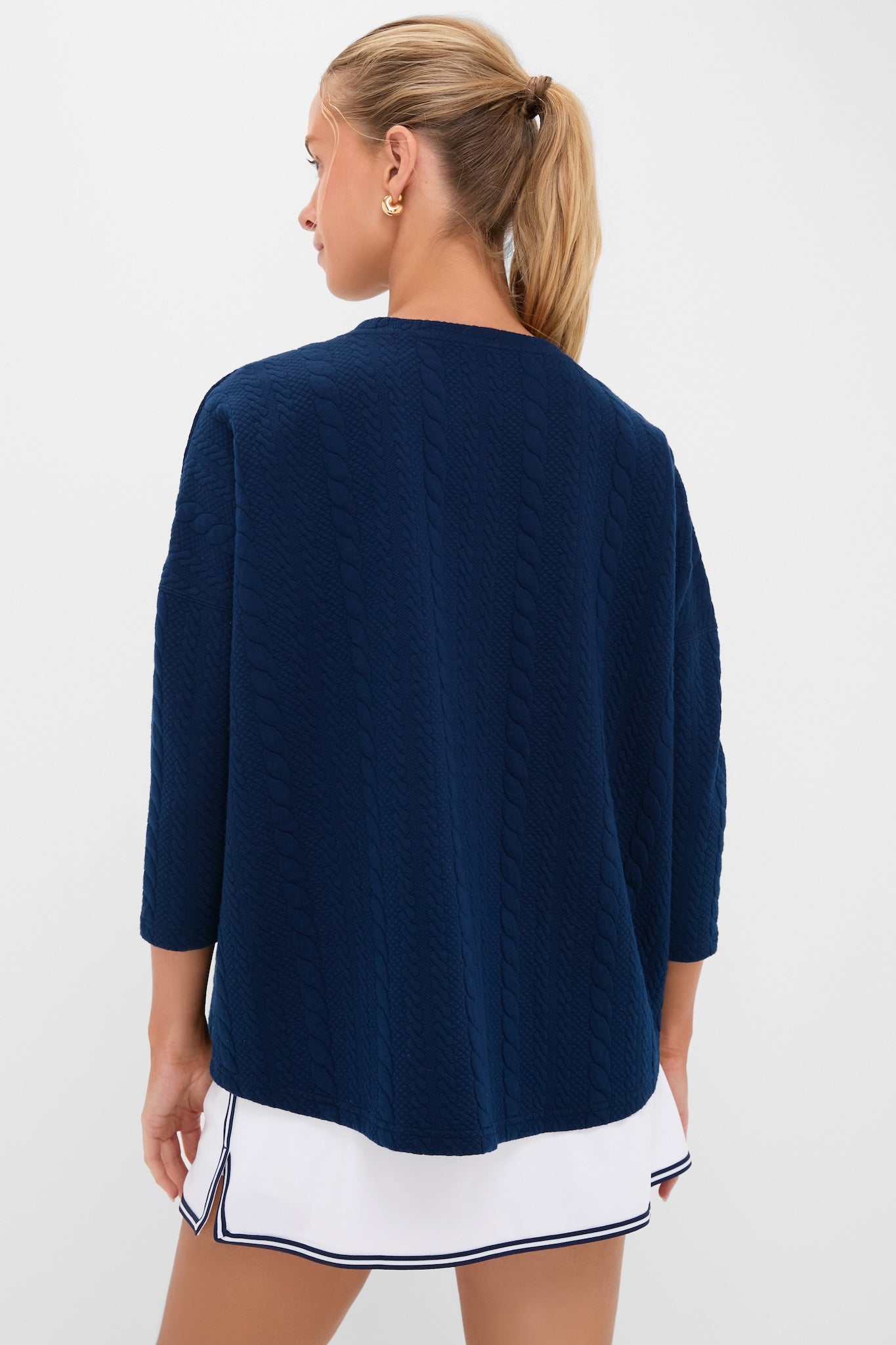Woman in a classic Navy Cable Ally Swing Sweatshirt with cable texture and three-quarter sleeves, paired effortlessly with a fun, high-end white skirt featuring navy trim.