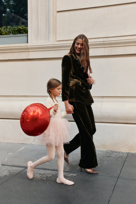 A woman wearing a dark olive, velvet matching blazer and pants set and heels walks with a little girl. The little girl wears a pink tutu, pink tights, pink ballet slippers, and a white cardigan while carrying a red balloon.