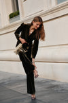 A woman wearing a dark olive velvet blazer, matching olive velvet pants, and t-strap heels holds a gold fringe purse.