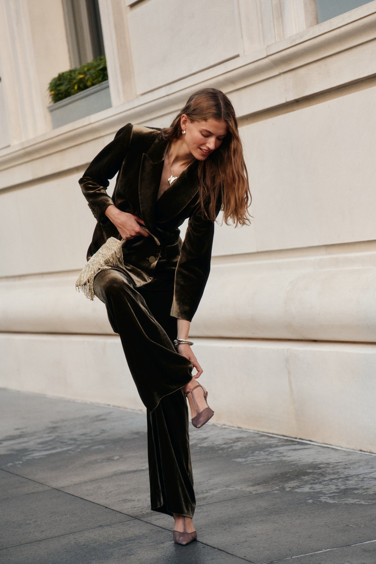 A woman wearing a dark olive velvet blazer, matching olive velvet pants, and t-strap heels holds a gold fringe purse.