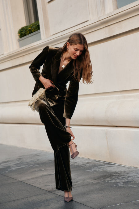 A woman wearing a dark olive velvet blazer, matching olive velvet pants, and t-strap heels holds a gold fringe purse.