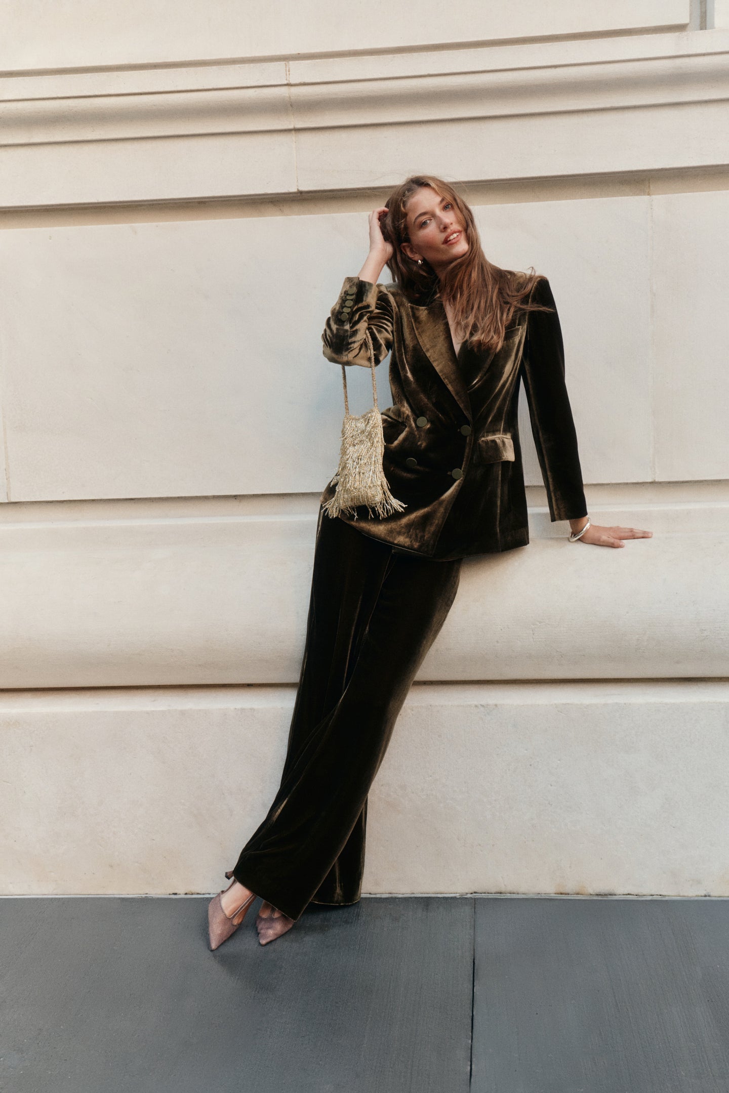 A woman wearing a dark olive velvet blazer, matching olive velvet pants, and t-strap heels holds a gold fringe purse.