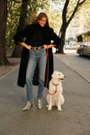 A woman walks a dog with a red leash while wearing a full-length fur coat, a black turtleneck, a pair of blue jeans, and a pair of snakeskin boots.